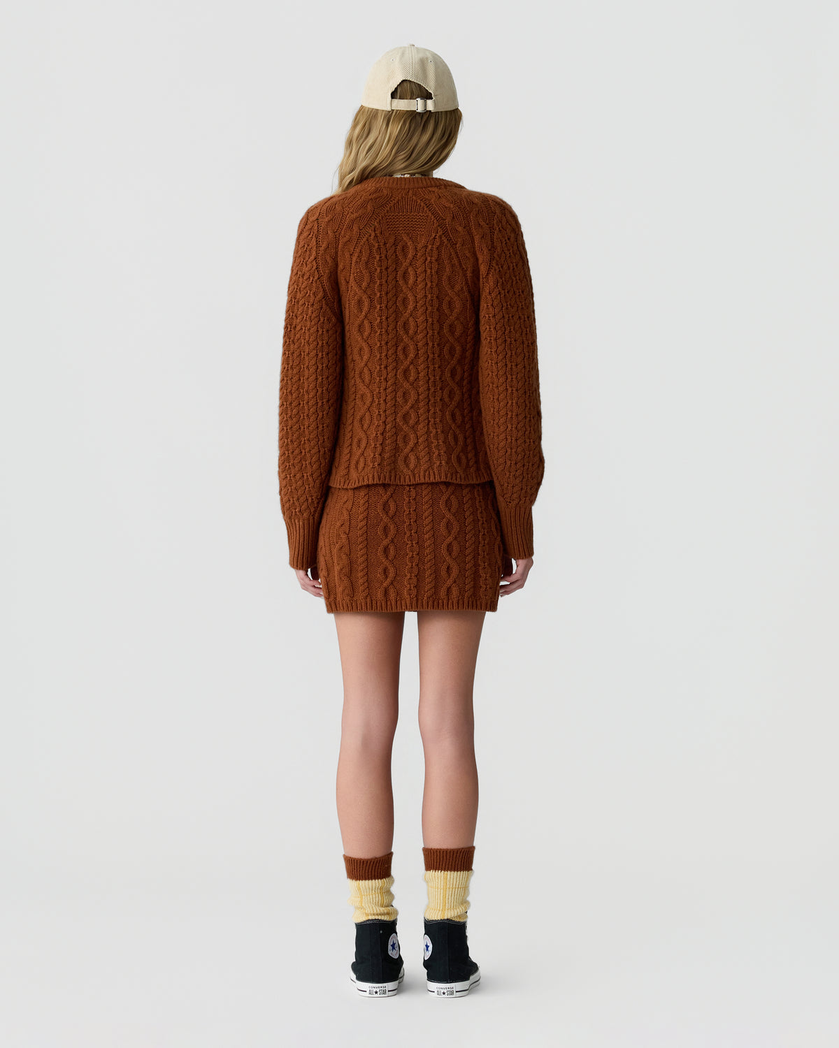 Back view of a model wearing a brown cable knit cardigan and matching mini skirt with a cream cap, striped socks, and black sneakers against a light background