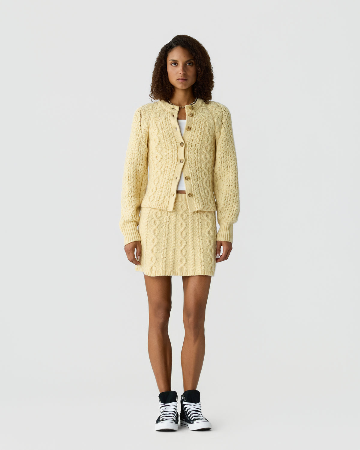 Woman wearing a cream-colored cable knit cardigan and matching skirt with black and white high-top sneakers, standing against a plain light background.