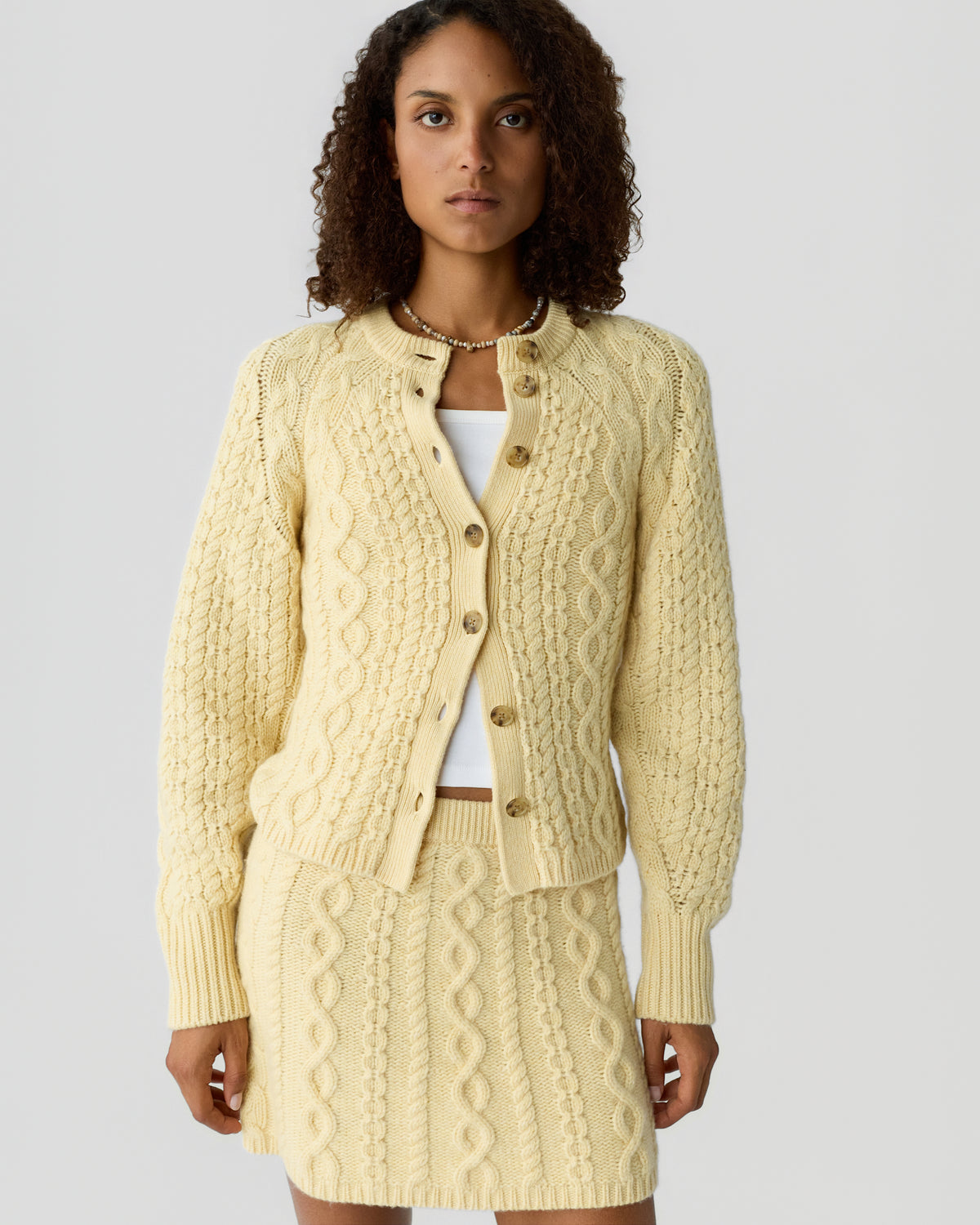Woman wearing a cream-colored cable knit cardigan with buttons and a matching cable knit skirt standing against a plain gray background.
