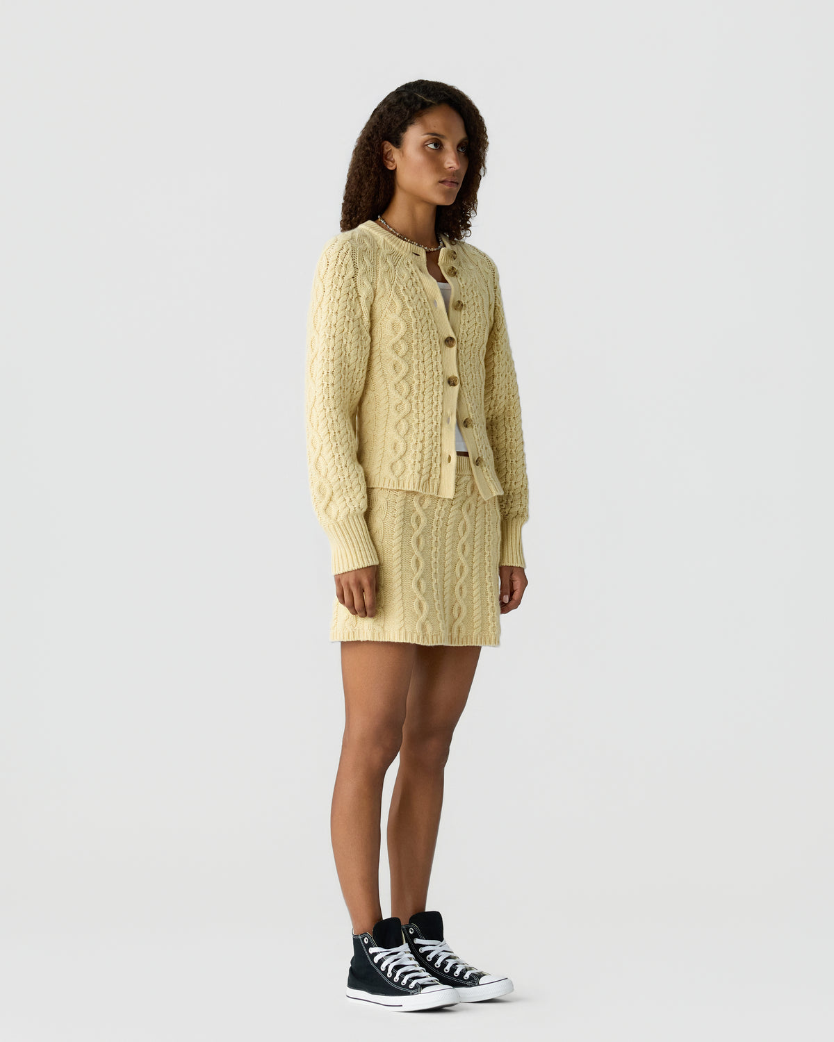 Woman wearing a cream-colored cable knit cardigan and matching skirt standing against a plain white background with black high-top sneakers.