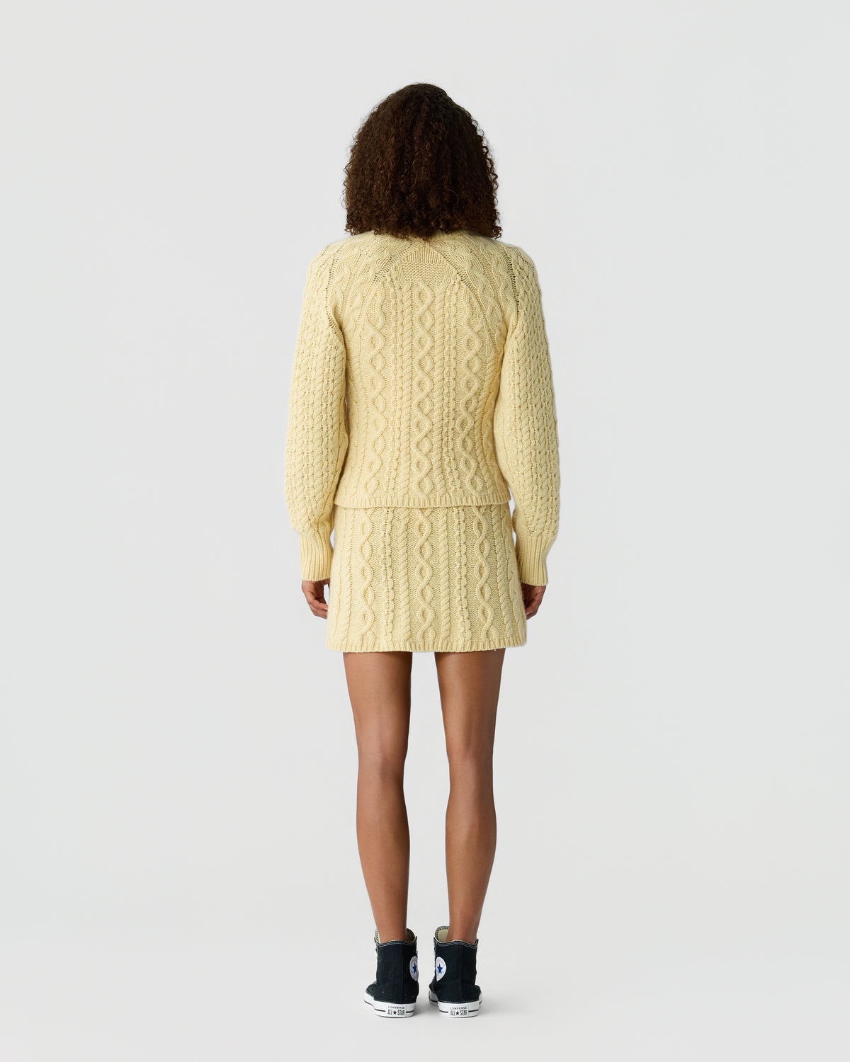 Woman seen from behind wearing a cream-colored cable knit sweater and matching knit skirt paired with black high-top sneakers against a plain background.
