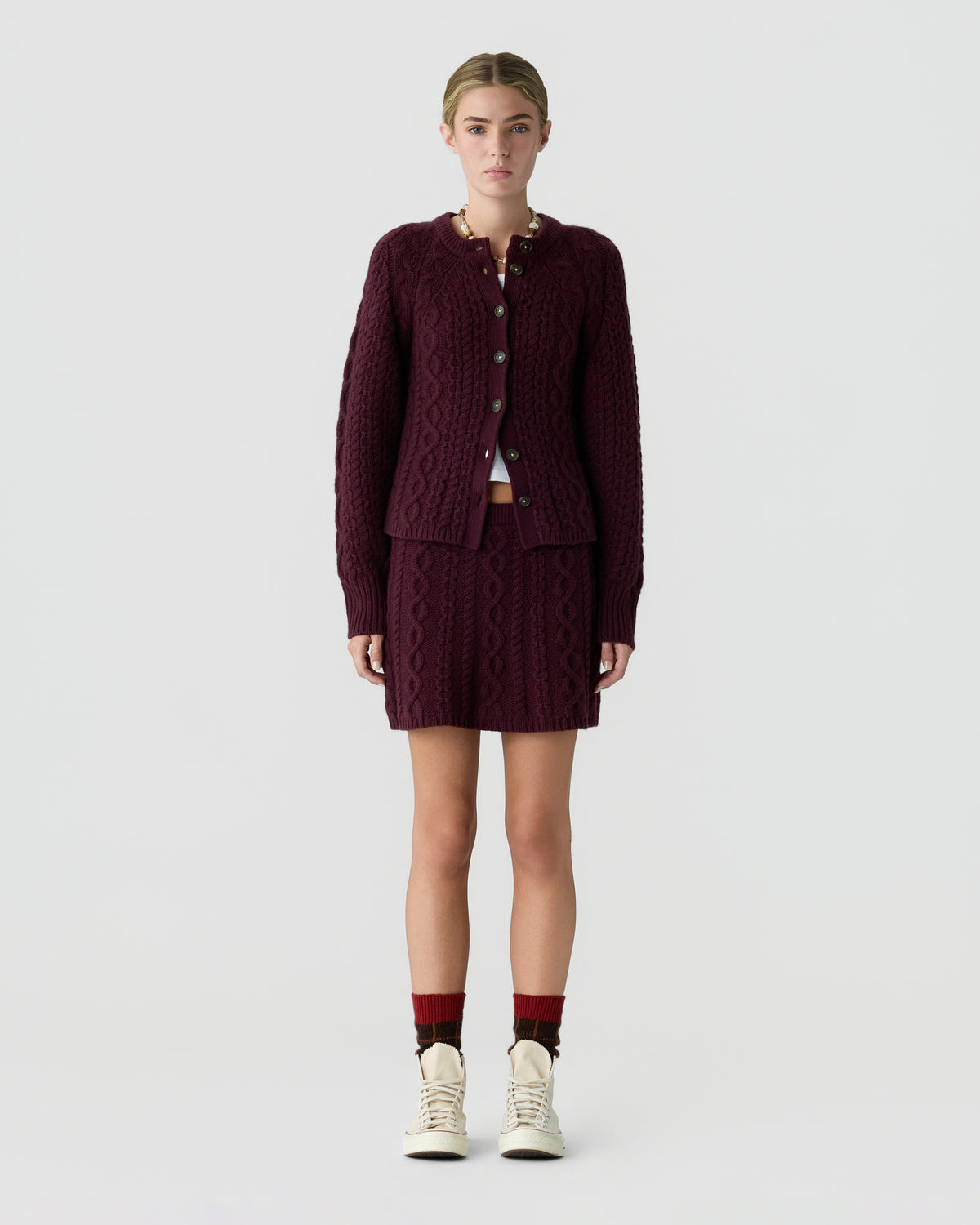 Model wearing a burgundy cable knit cardigan with matching skirt, burgundy socks, and white sneakers against a light gray background