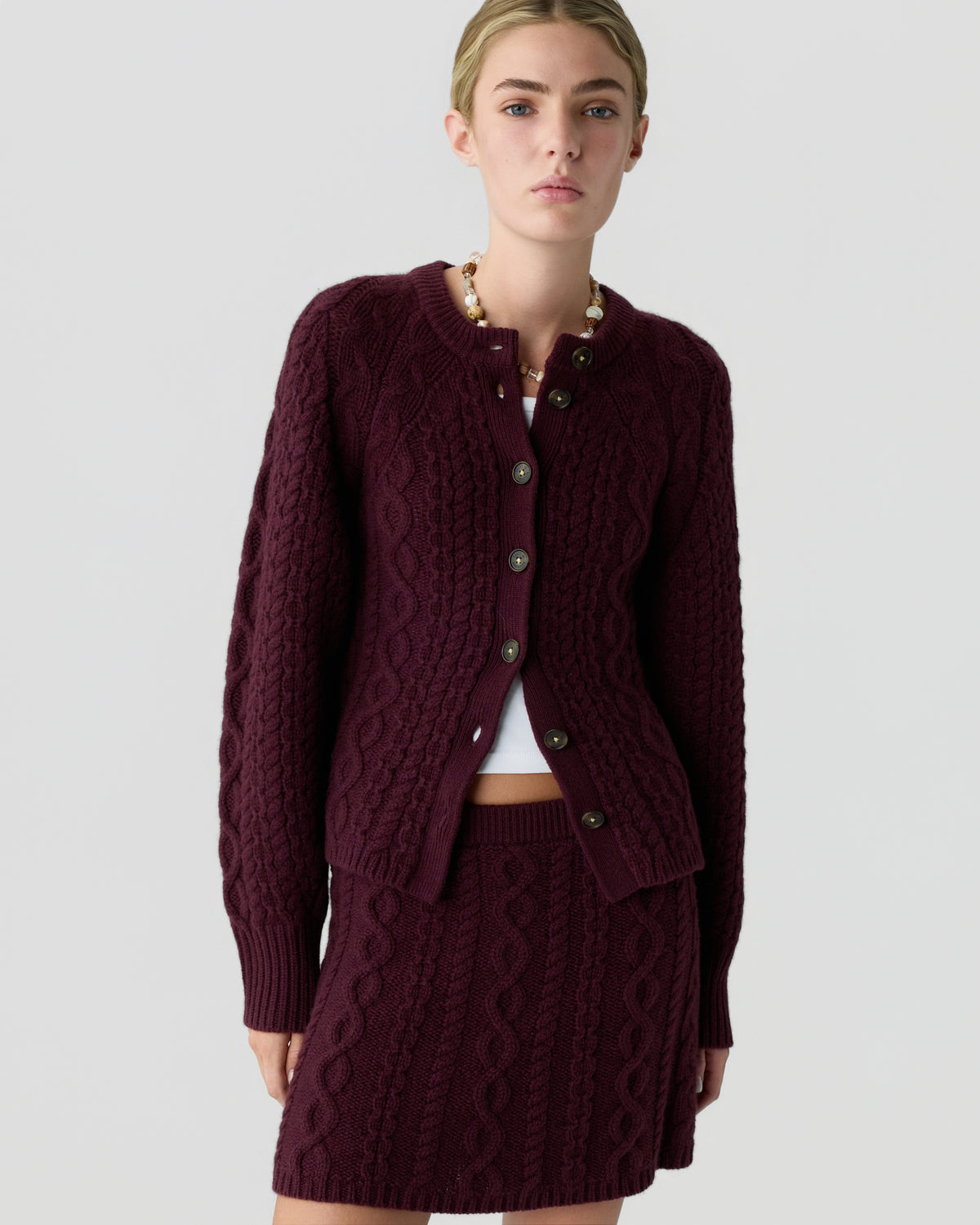 Model wearing a burgundy cable knit button front cardigan with a matching textured mini skirt and beaded necklace against a light gray background