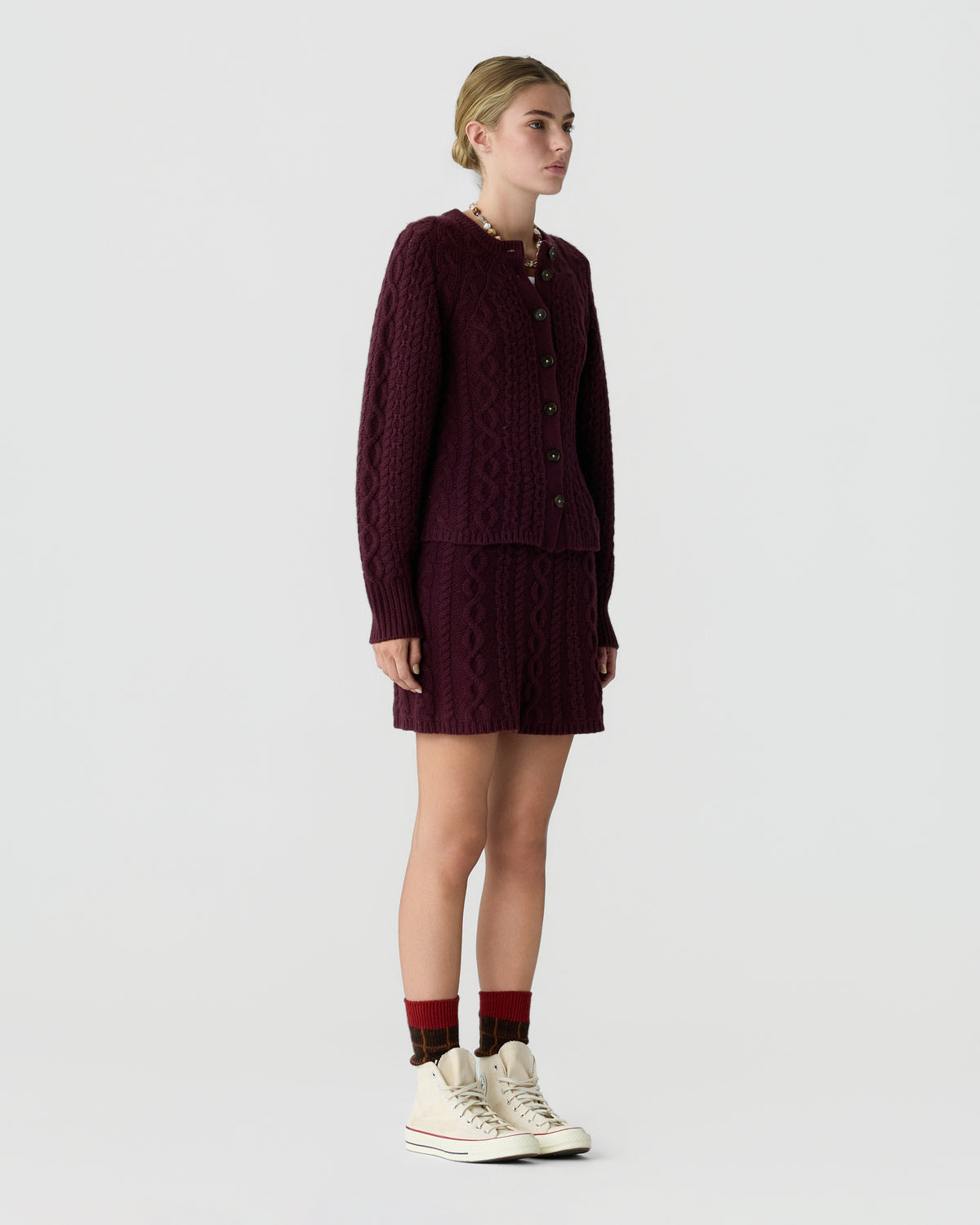 A model wears a burgundy cable knit cardigan and matching pleated skirt with red socks and cream high top sneakers against a light gray background