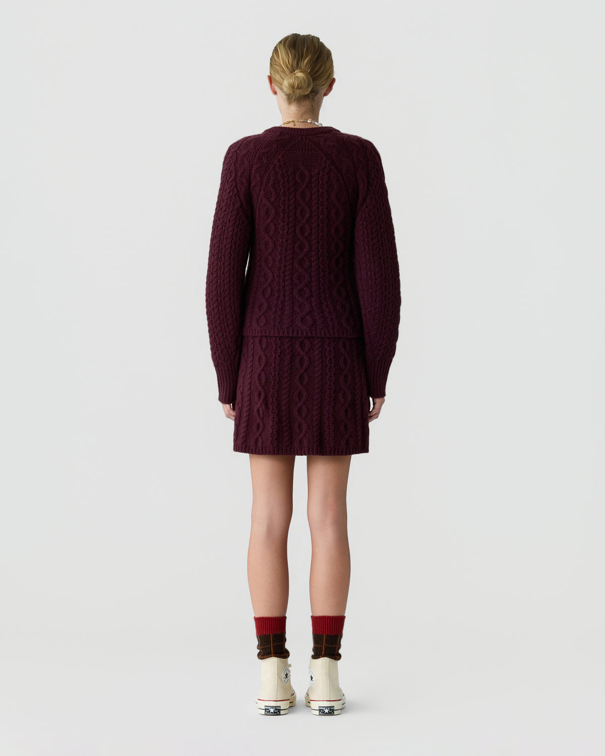 Back view of a model wearing a burgundy cable knit cardigan and matching short skirt with dark socks and white sneakers against a light gray background