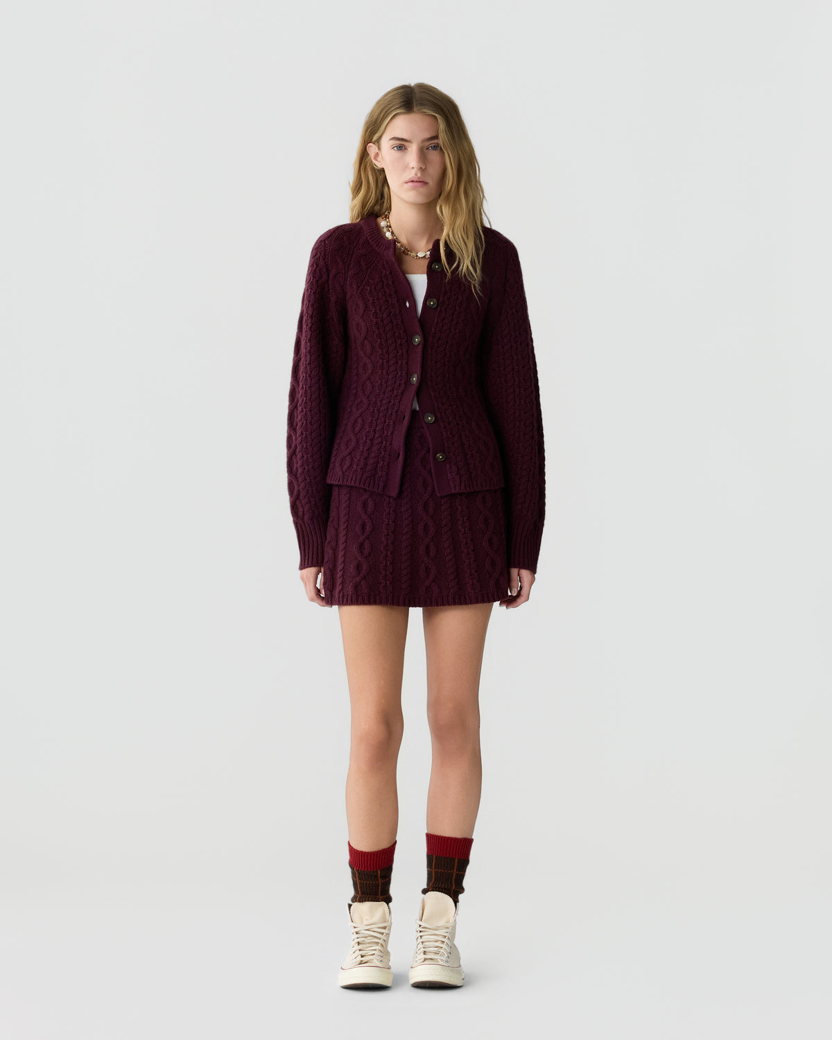 Young woman wearing a dark purple cable-knit cardigan and matching skirt, paired with cream lace-up shoes and patterned socks, standing against a plain white background.