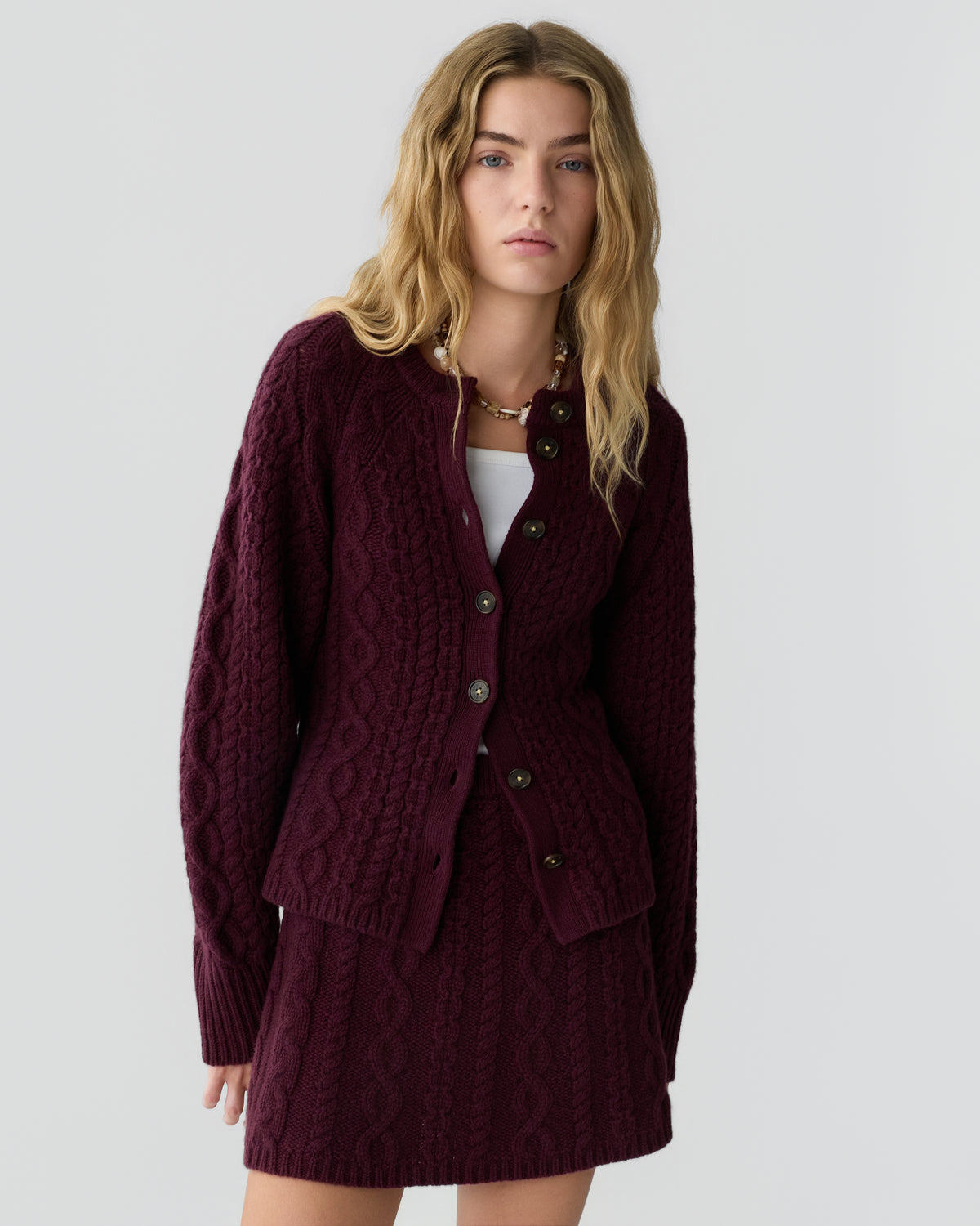 Woman wearing a matching deep burgundy cable knit cardigan and skirt set, standing against a plain light gray background with a neutral expression.