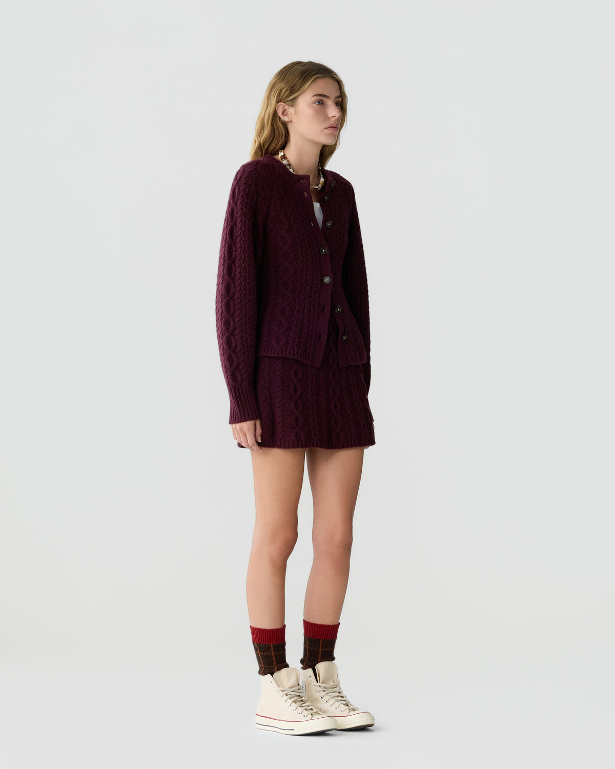 Young woman wearing a burgundy cable knit cardigan and matching skirt standing against a plain light background with white high-top sneakers and patterned socks.