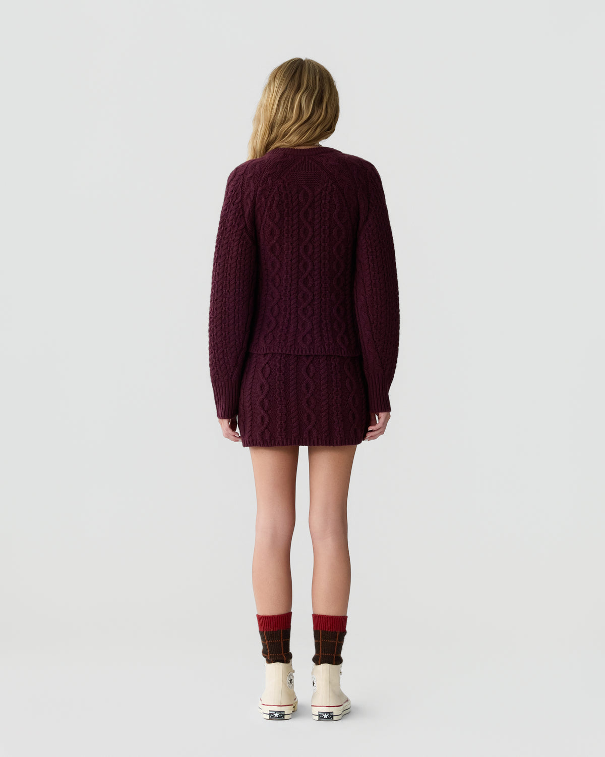 A person with long blonde hair stands facing away, wearing a dark purple cable-knit sweater and matching mini skirt, paired with patterned socks and light-colored high-top sneakers.