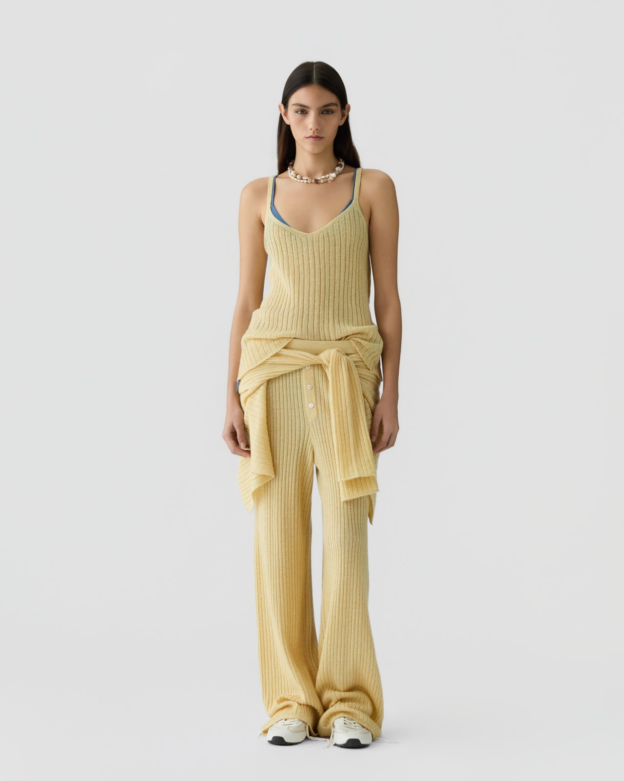 Woman standing facing forward wearing a ribbed yellow sleeveless tank top layered over a blue camisole, paired with matching wide-leg ribbed pants and a tied long-sleeve sweater around the waist with white sneakers.