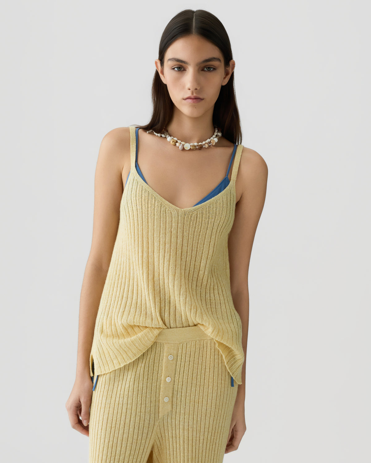 Young woman wearing a ribbed light yellow sleeveless knit top layered over a blue camisole, paired with matching ribbed pants and a shell necklace against a plain background.