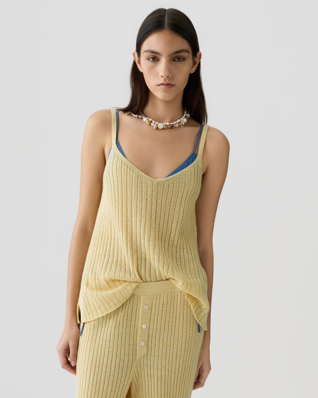 Young woman wearing a ribbed light yellow sleeveless knit top layered over a blue camisole, paired with matching ribbed pants and a shell necklace against a plain background.