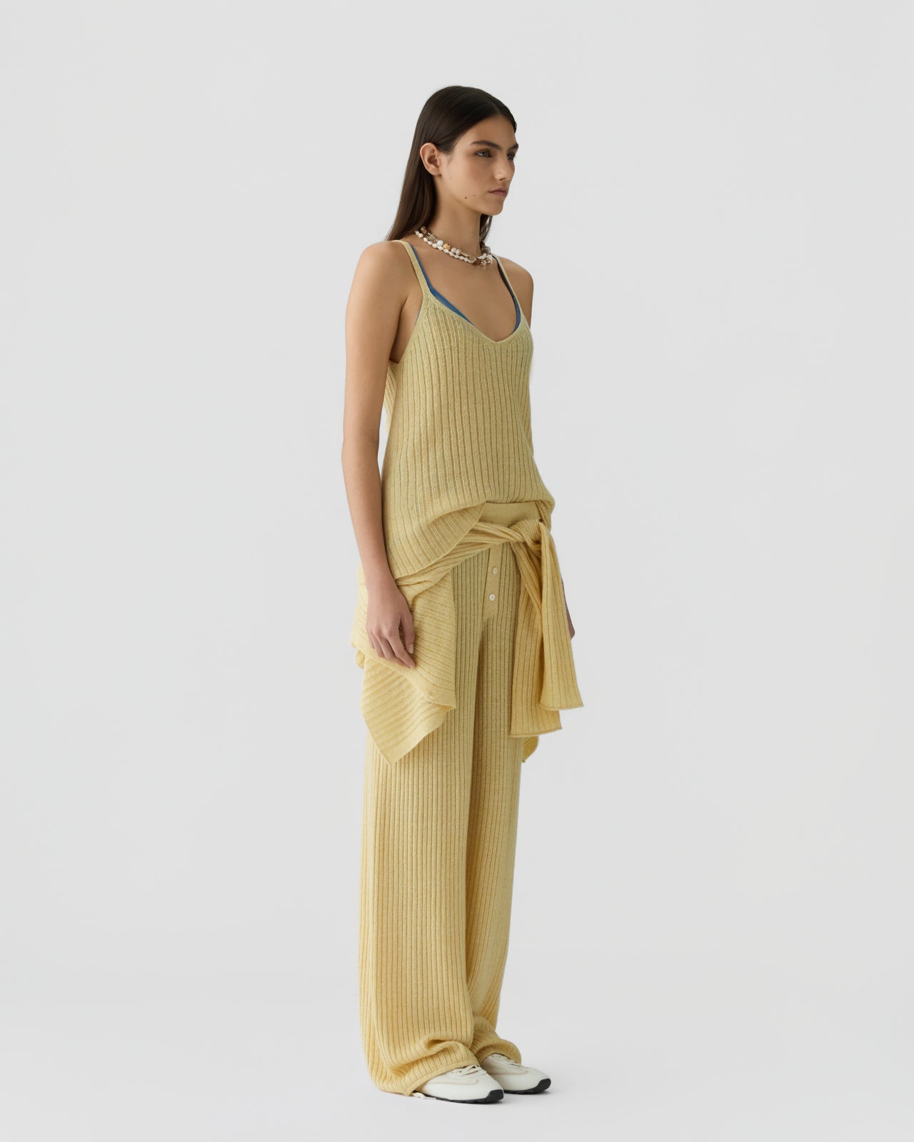 Woman standing in profile wearing a soft yellow ribbed sleeveless top layered over a blue camisole, paired with matching wide-leg ribbed pants and a tied knit sweater around the waist.