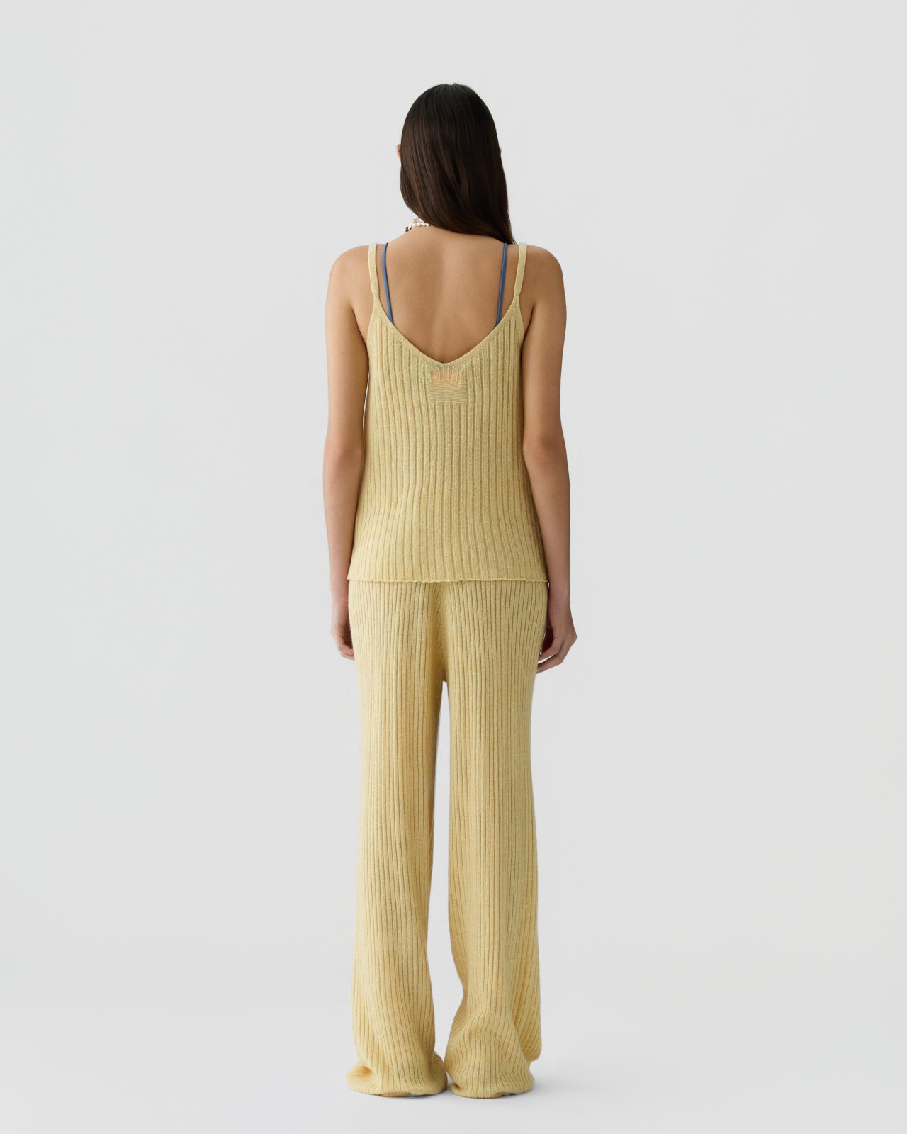 Woman shown from behind wearing a ribbed light yellow sleeveless knit top with thin straps, paired with matching wide-leg ribbed pants against a plain white background.