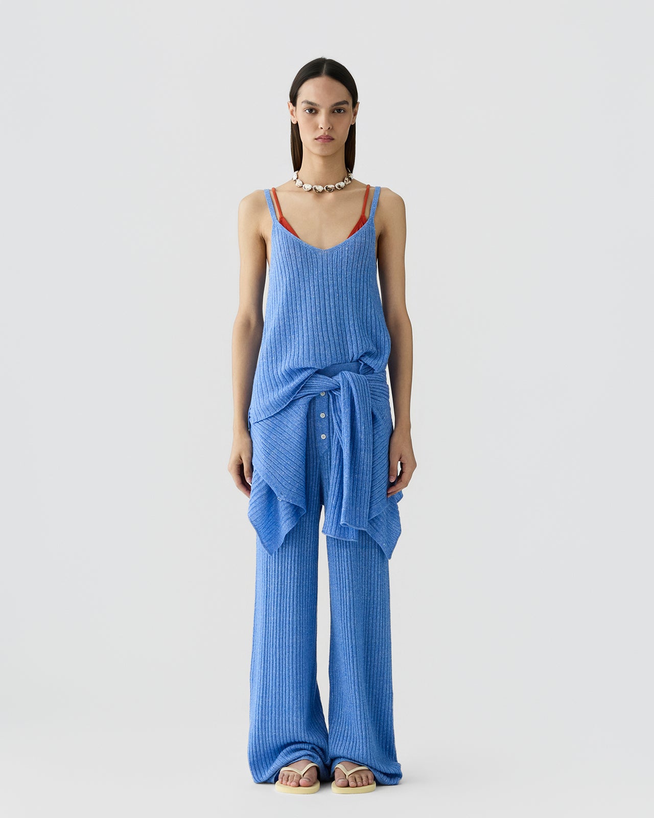 Woman facing forward wearing a ribbed sleeveless blue tank top layered over a red camisole, paired with matching wide-leg ribbed pants and a tied long-sleeve sweater around the waist.