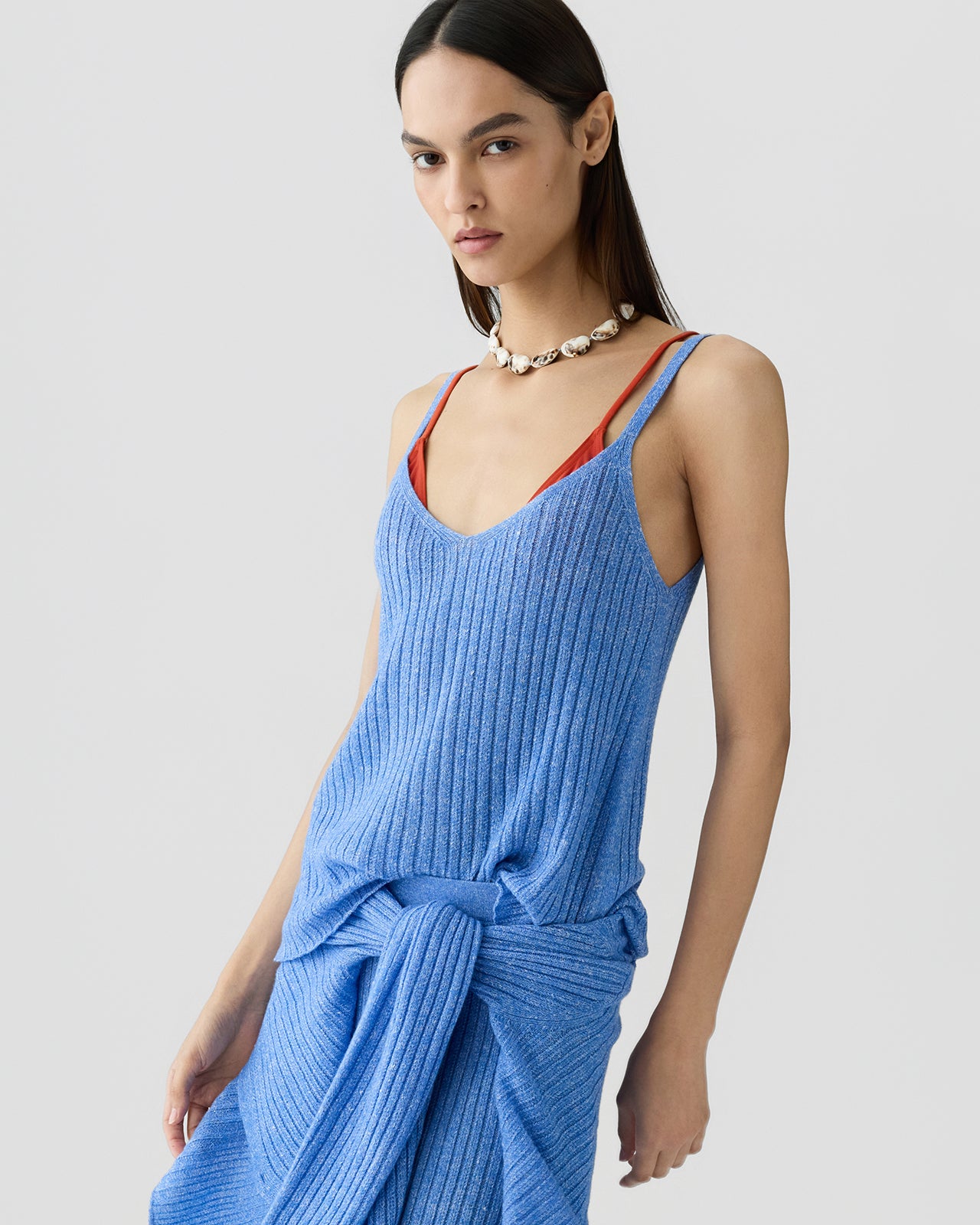 Woman facing forward wearing a ribbed blue sleeveless tank top layered over a red camisole, styled with a matching ribbed skirt tied at the waist and a shell necklace.
