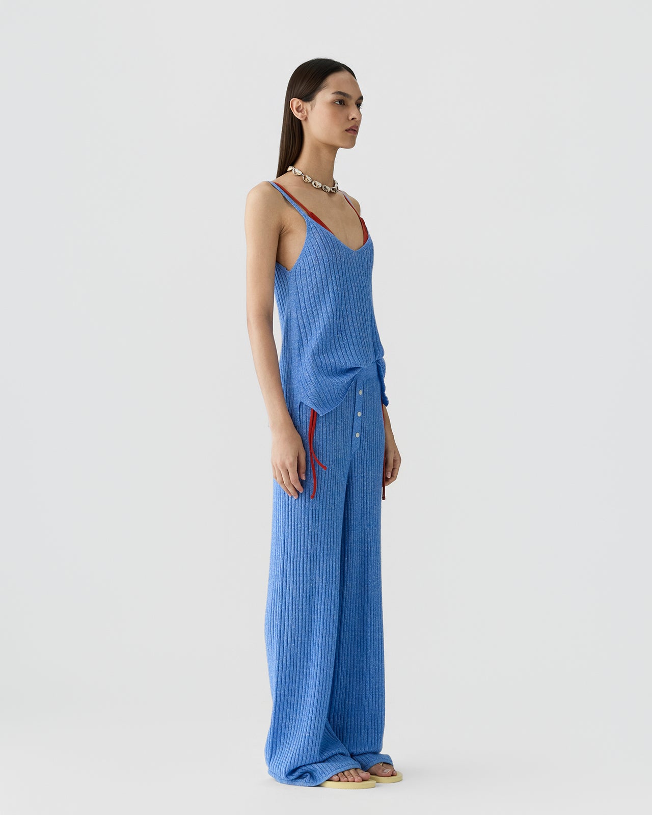 Woman standing in profile wearing a sleeveless ribbed blue tank top with red straps layered over a red bra, paired with matching wide-leg ribbed pants and barefoot.