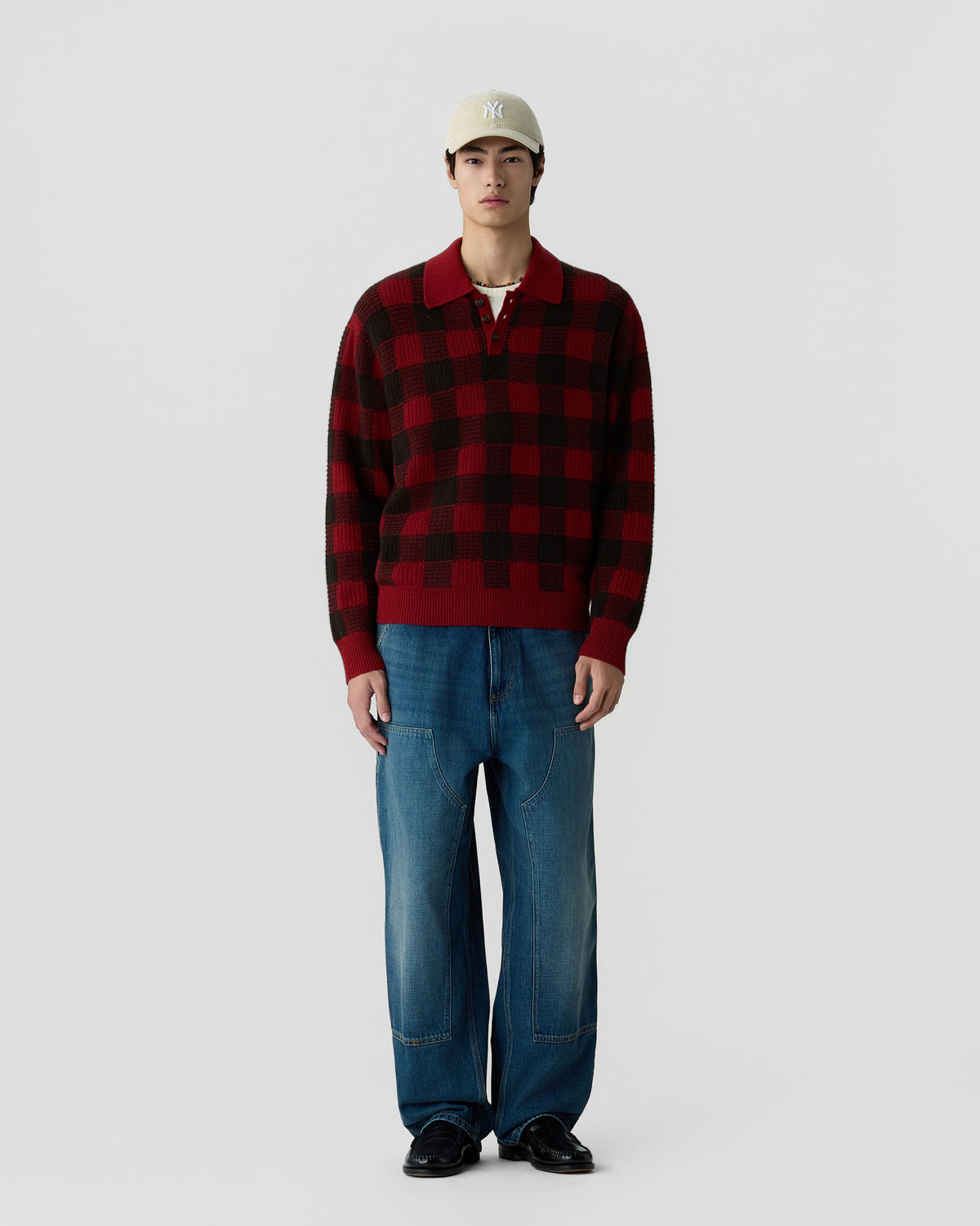 A young man wearing a beige baseball cap, a red and black checkered pullover sweater, loose-fitting blue jeans, and black shoes standing against a plain white background.