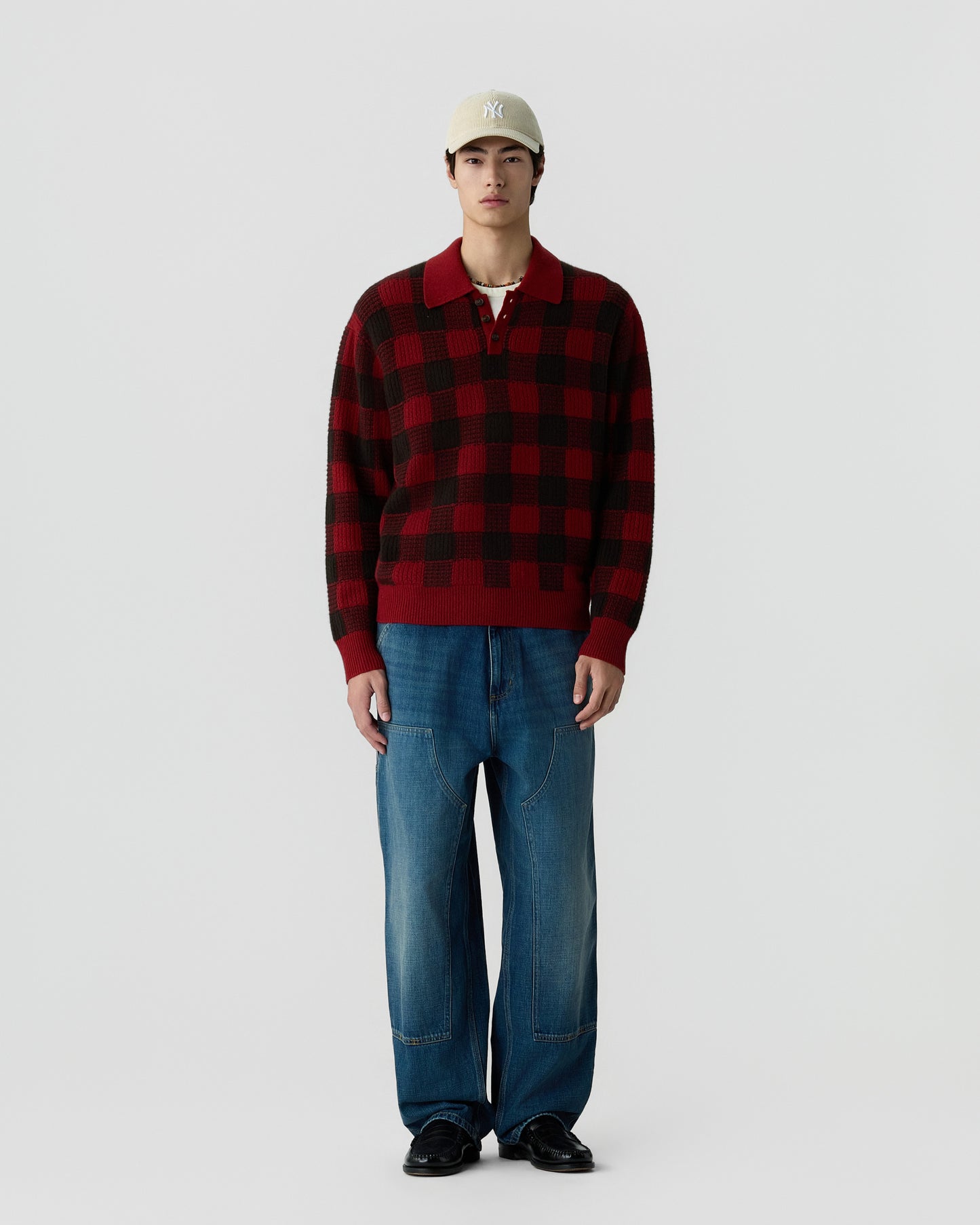 A young man wearing a beige baseball cap, a red and black checkered pullover sweater, loose-fitting blue jeans, and black shoes standing against a plain white background.
