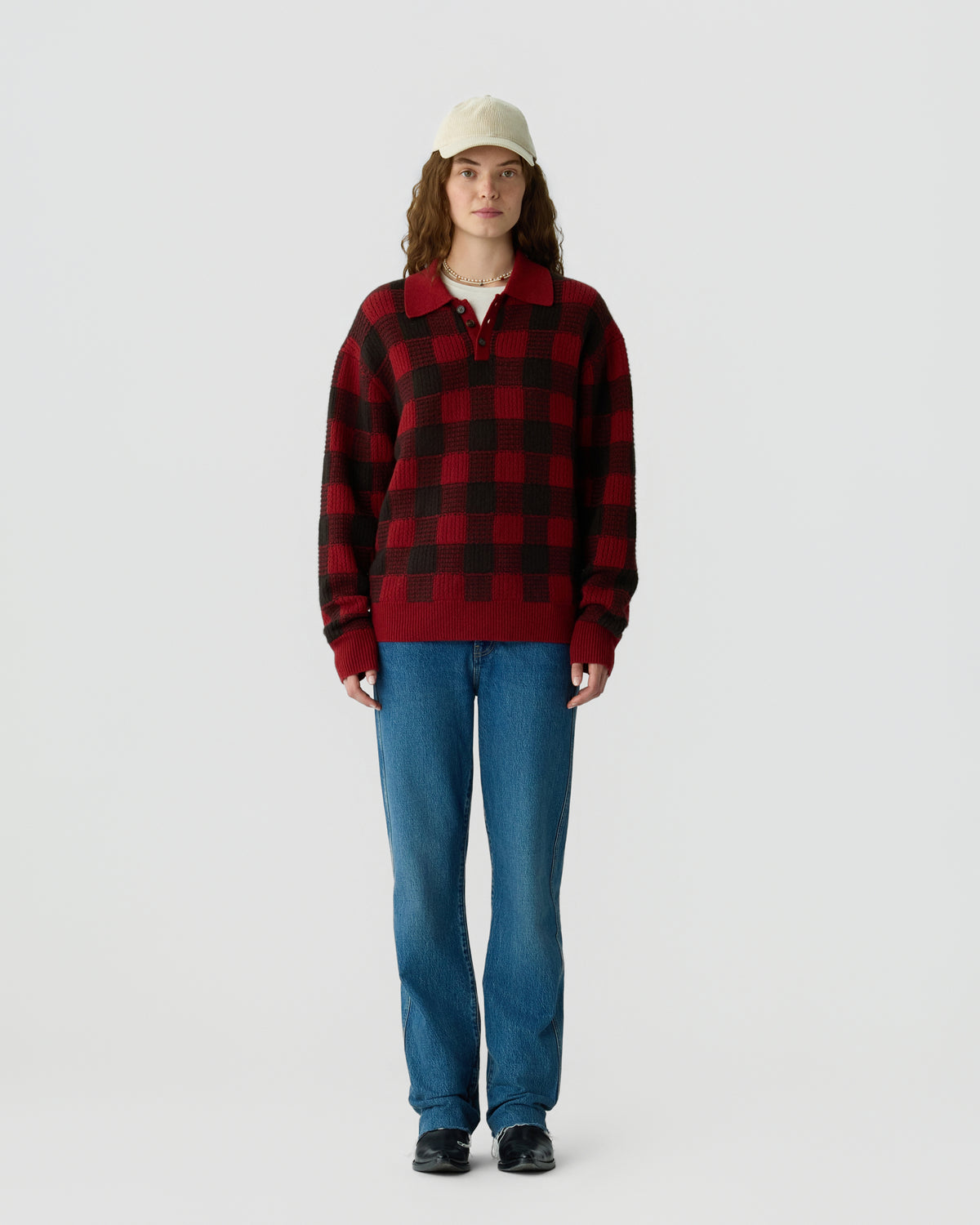 Young woman wearing a red and black checkered sweater, blue jeans, black shoes, and a beige baseball cap, standing against a plain light background.