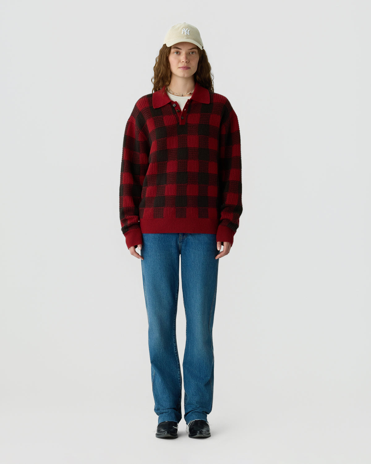 Full-length person wearing a red and black checkered pullover with blue jeans black shoes and a light beige baseball cap against a pale gray background
