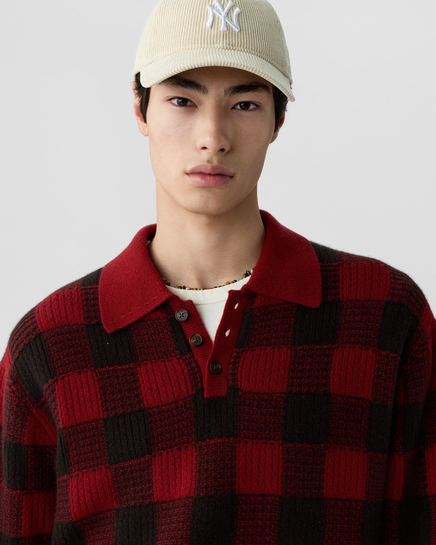 Close-up of a young man wearing a beige baseball cap and a red and black checkered knit sweater with a collar and buttons against a plain light background.