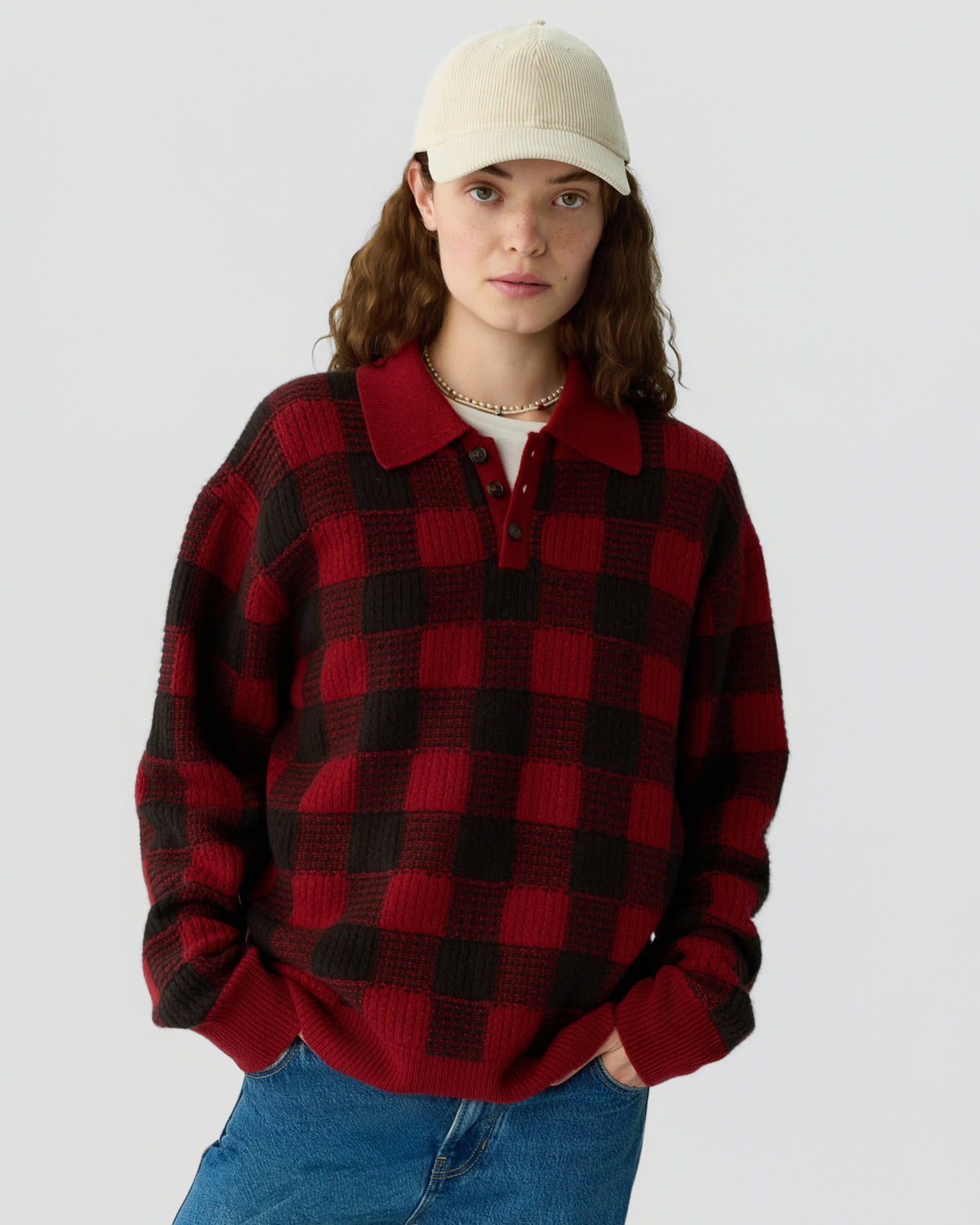 Young woman wearing a red and black checkered knit sweater with a collar and buttons, paired with blue jeans and an off-white baseball cap against a plain background.