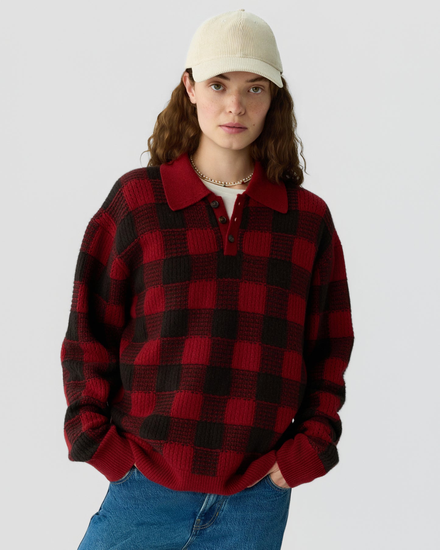 Young woman wearing a red and black checkered knit sweater with a collar and buttons, paired with blue jeans and an off-white baseball cap against a plain background.