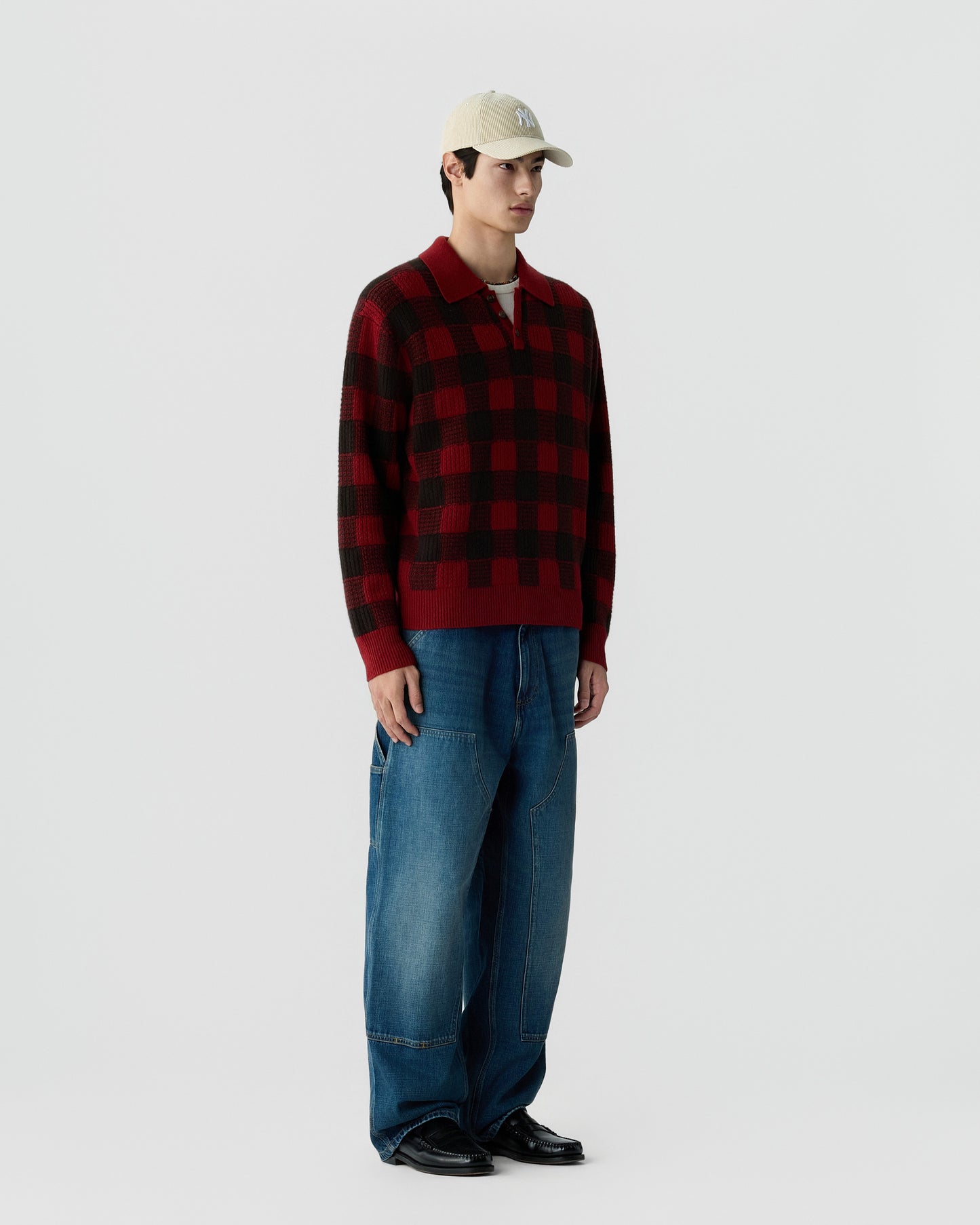 A man wearing a red and black checkered sweater, loose blue jeans, black shoes, and a light beige baseball cap standing against a plain white background.