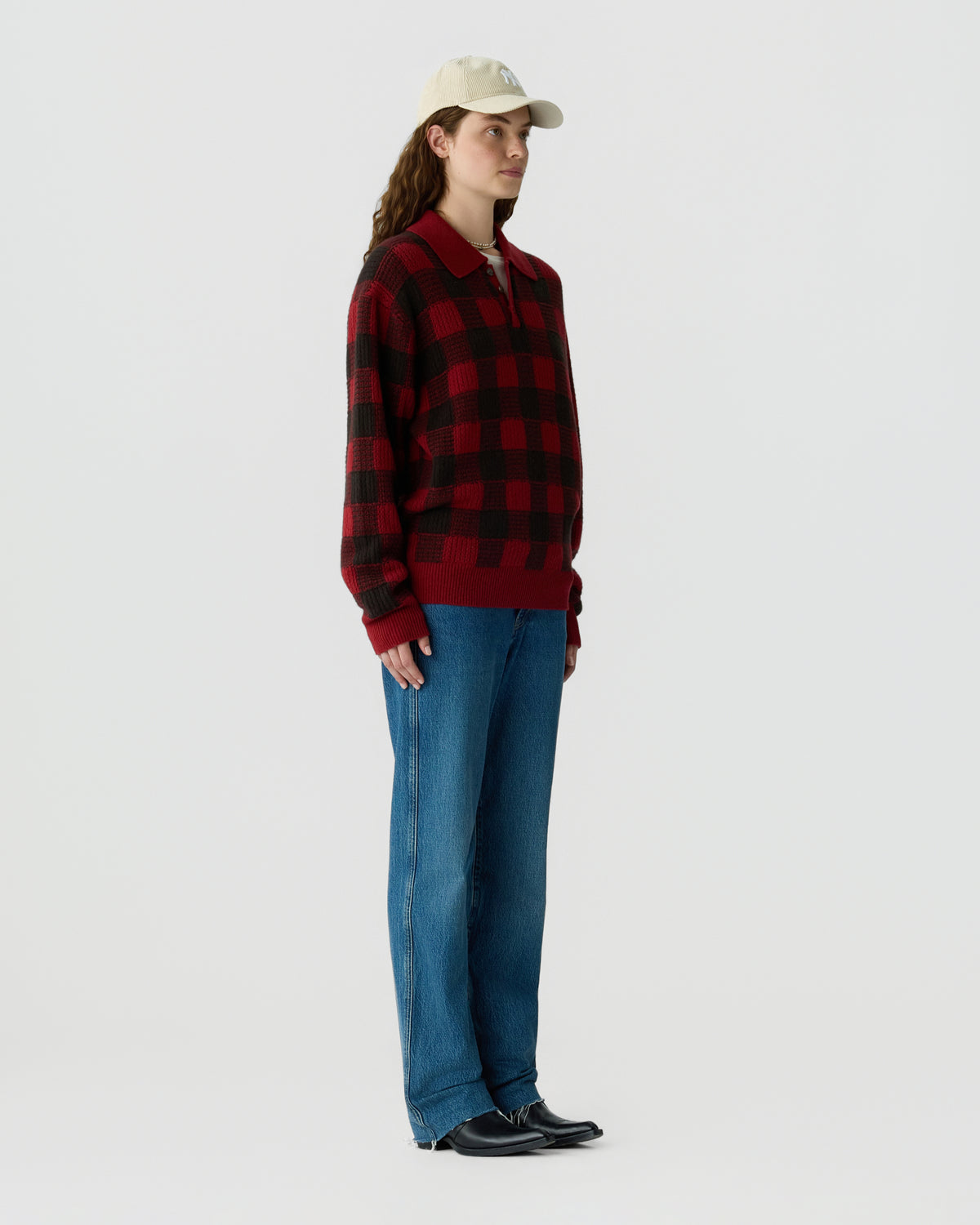 Woman wearing a red and black plaid sweater with blue jeans a beige cap and black shoes standing against a light gray background