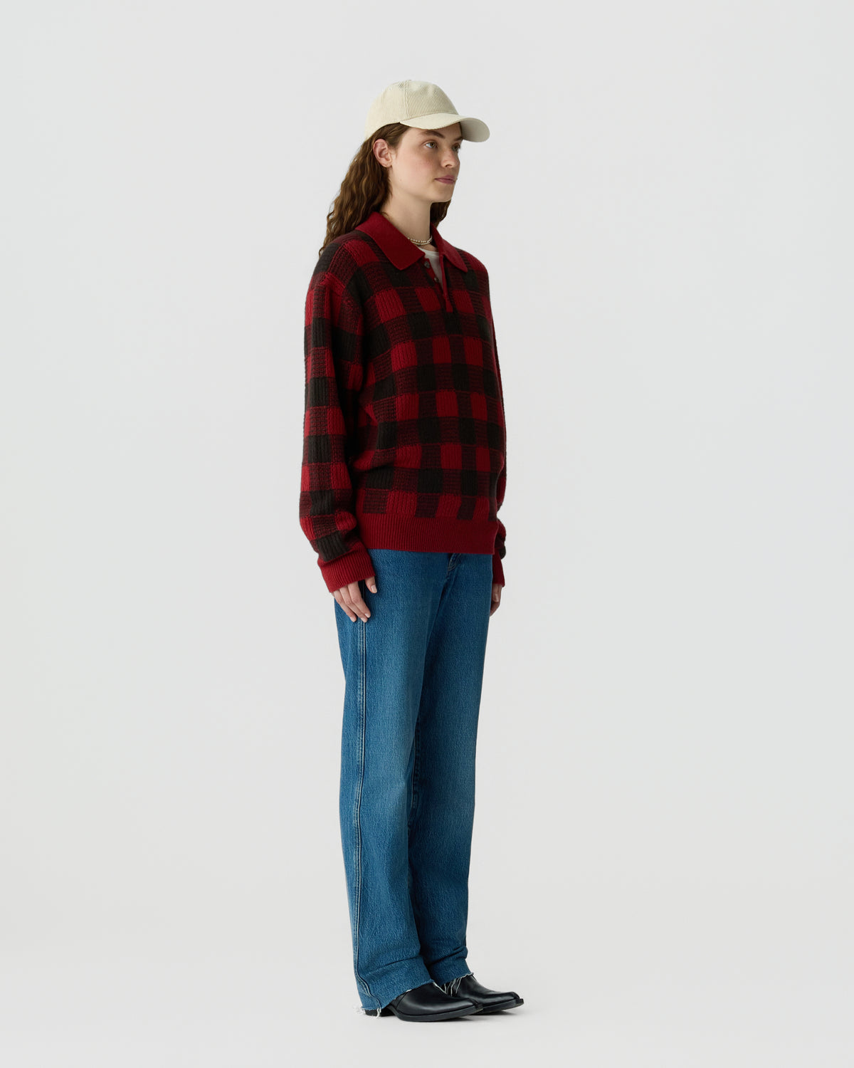 A person wearing a red and black checkered long-sleeve sweater, blue jeans, black shoes, and a beige baseball cap standing against a plain light gray background.