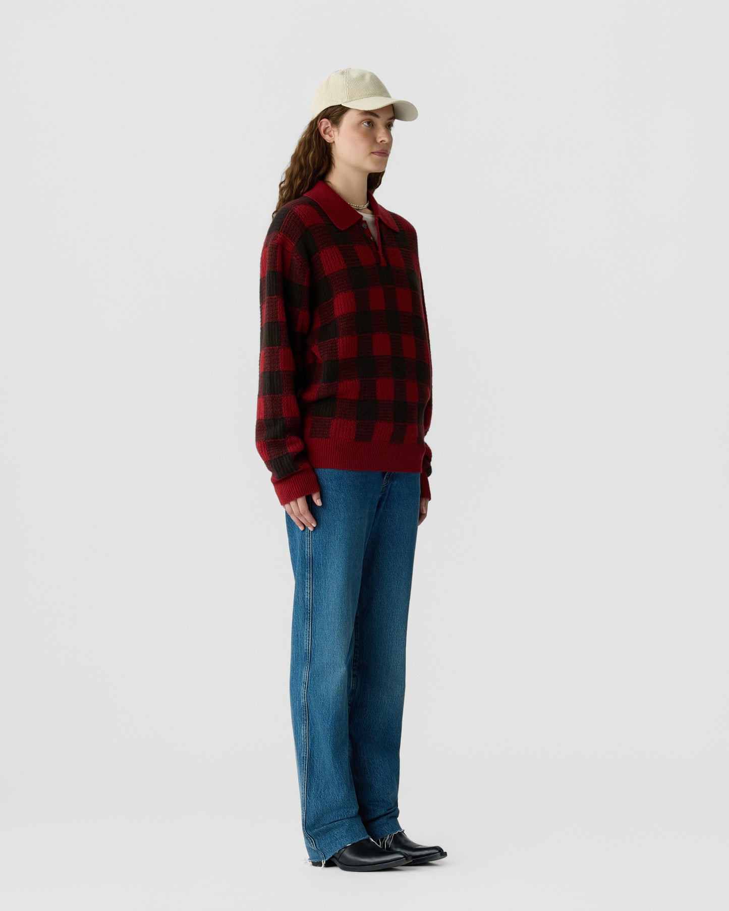 A person wearing a red and black checkered long-sleeve sweater, blue jeans, black shoes, and a beige baseball cap standing against a plain light gray background.