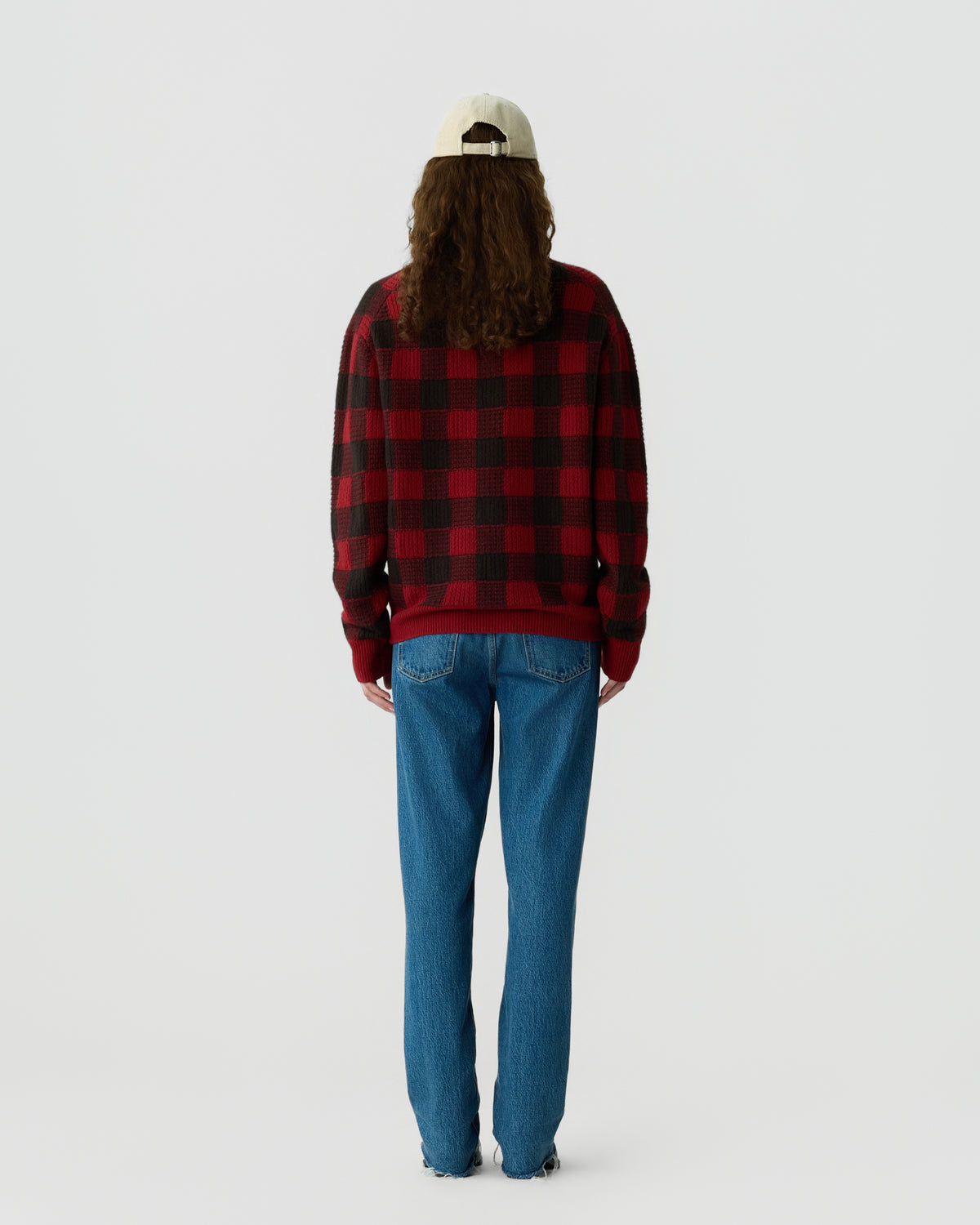 Person with long hair wearing a beige cap, red and black checkered sweater, and blue jeans standing facing away against a plain white background.