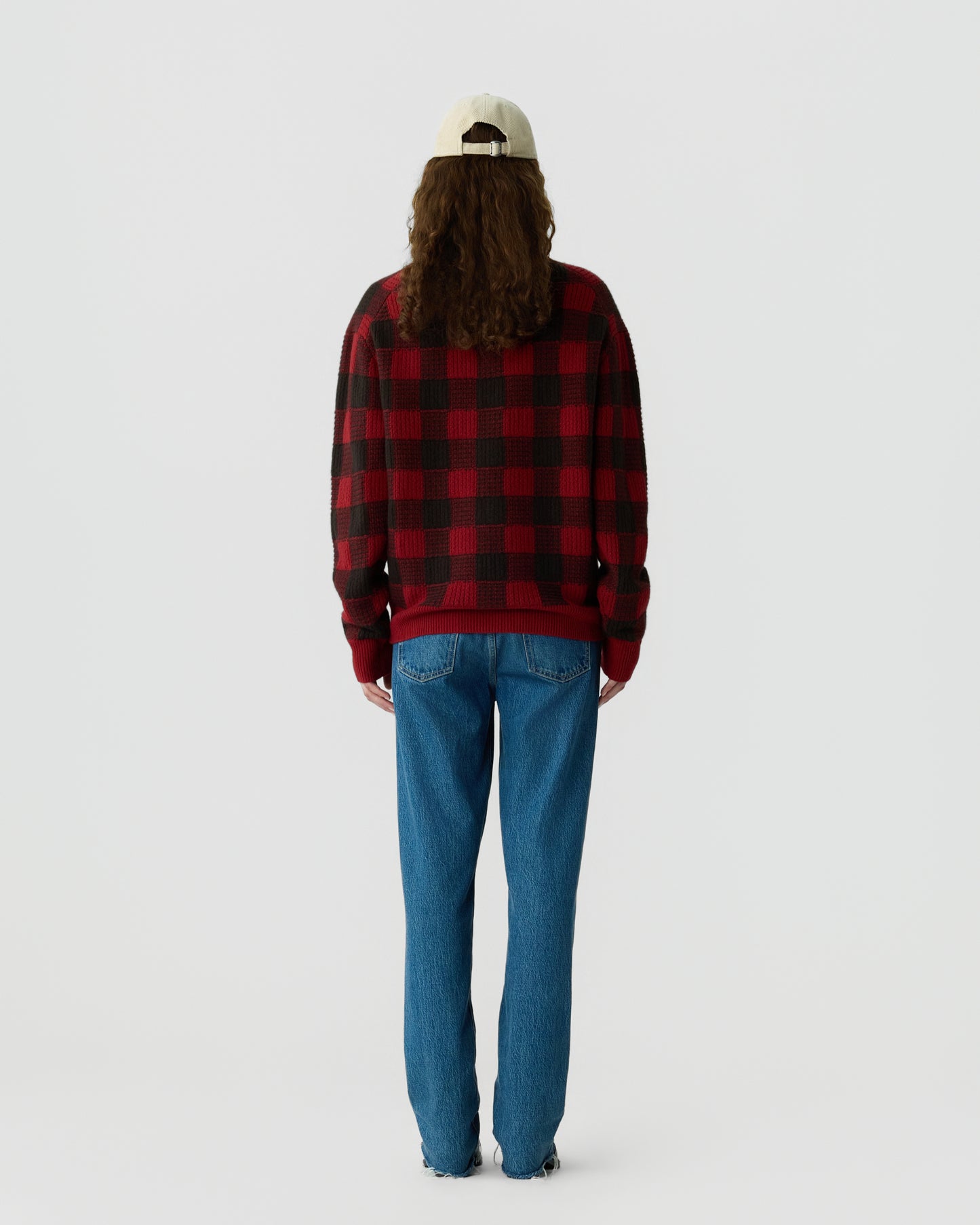 Person with long hair wearing a beige cap, red and black checkered sweater, and blue jeans standing facing away against a plain white background.