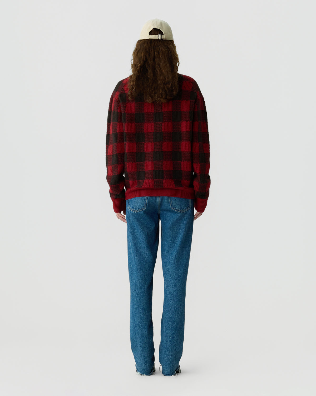 Back view of a person wearing a red and black plaid sweater with blue jeans and a light colored baseball cap against a plain white background