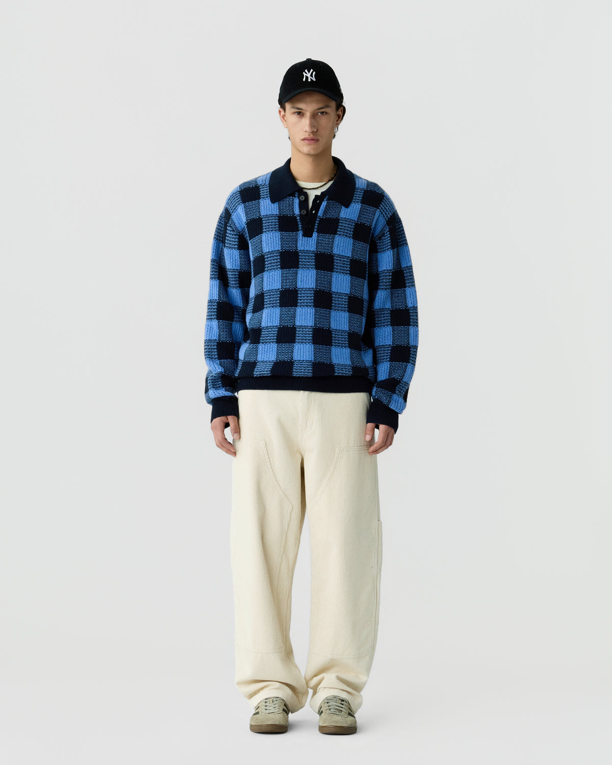 A young man wearing a blue and black checkered long-sleeve sweater, cream-colored wide-leg pants, beige shoes, and a black baseball cap with a white logo, standing against a plain white background.