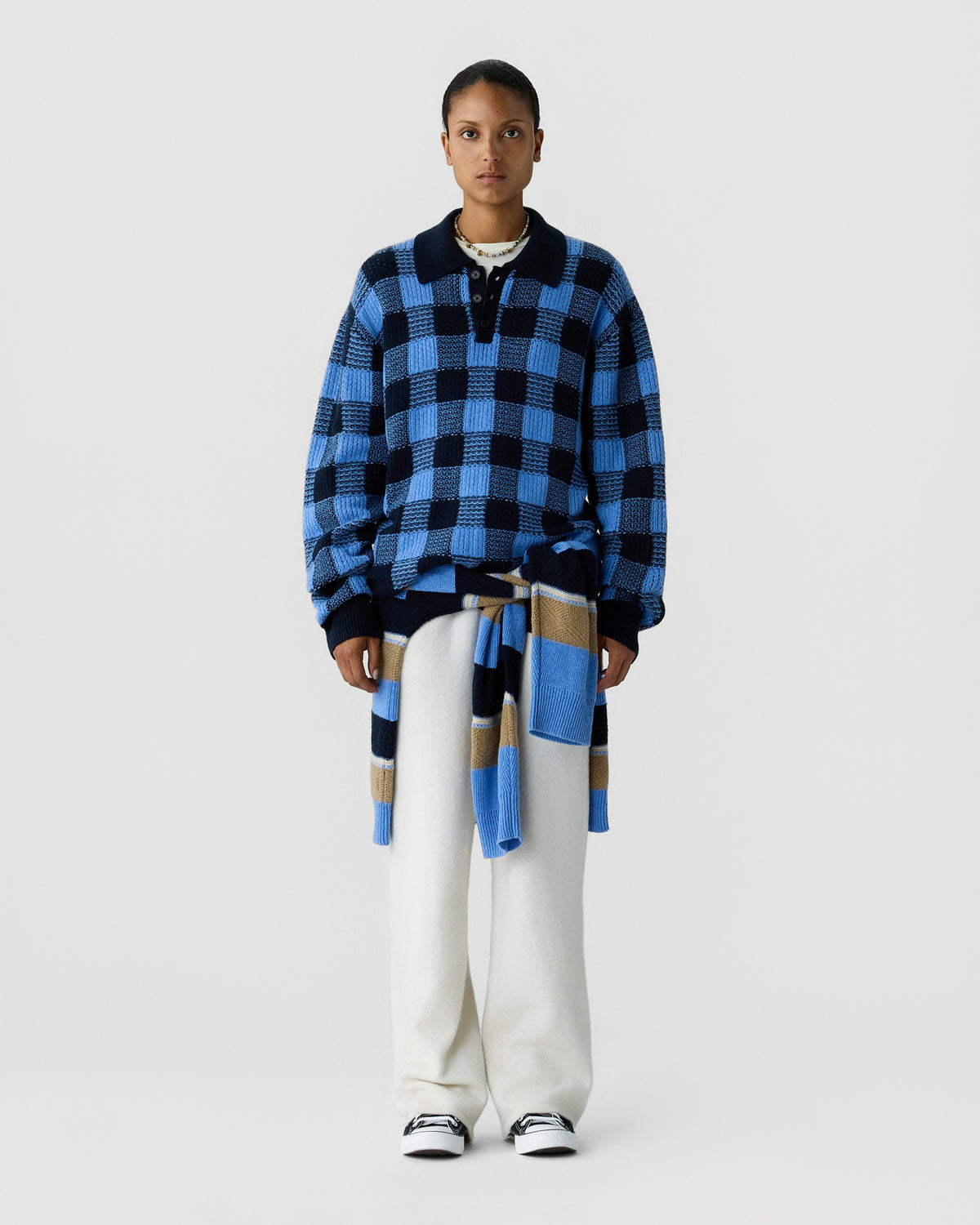 Person wearing a blue and black checkered sweater with a coordinating scarf tied at the waist white trousers and black and white sneakers against a light gray background
