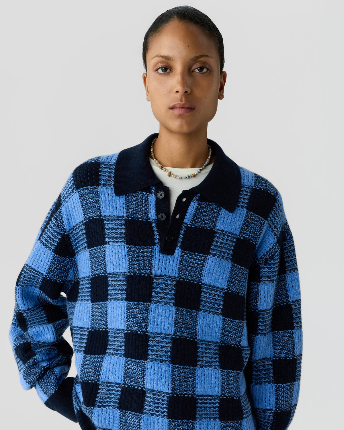 A woman wearing a blue and black checkered knit sweater with a black collar and button placket, standing against a plain light gray background.