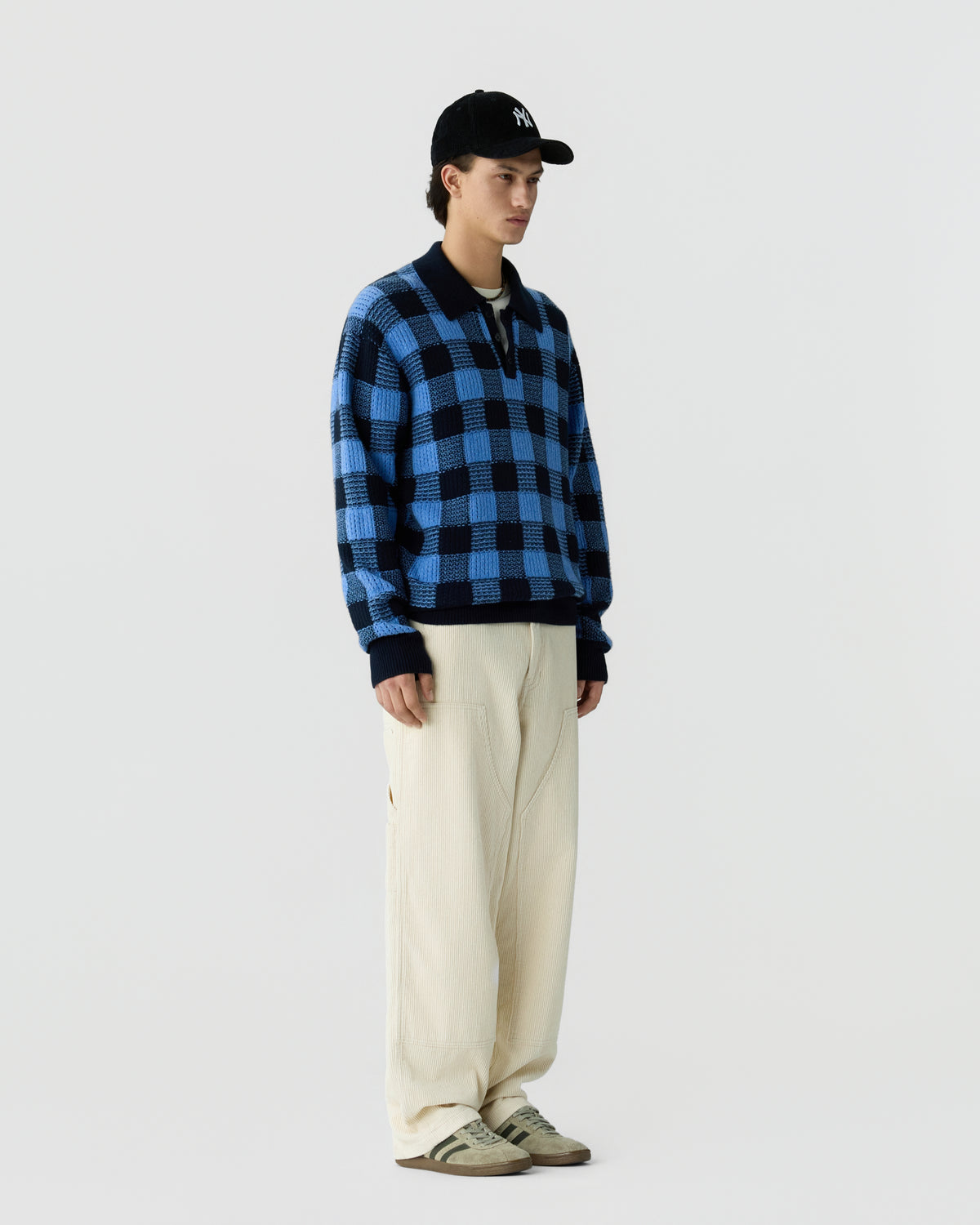 Young man wearing a blue and black checkered pullover sweater, cream wide-leg pants, beige sneakers, and a black baseball cap against a plain white background.