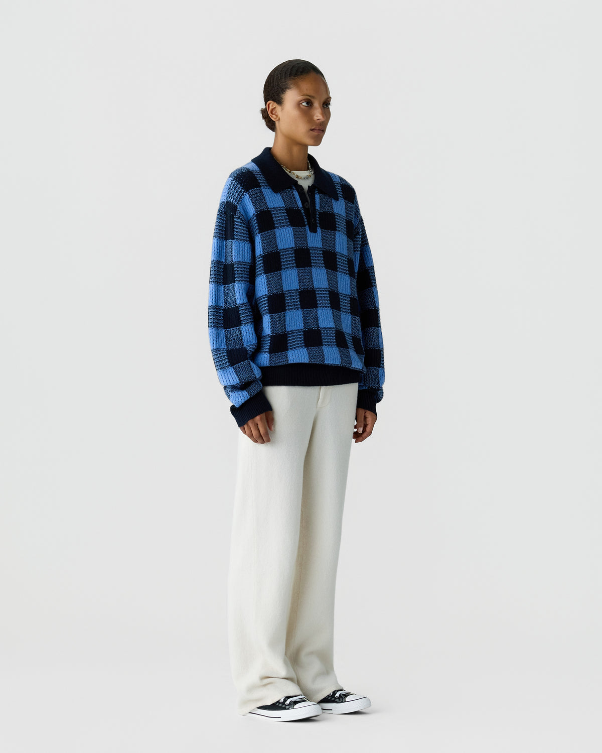 A person wearing a blue and black checkered long-sleeve pullover with a collar, white wide-leg pants, and black and white sneakers, standing against a plain background.