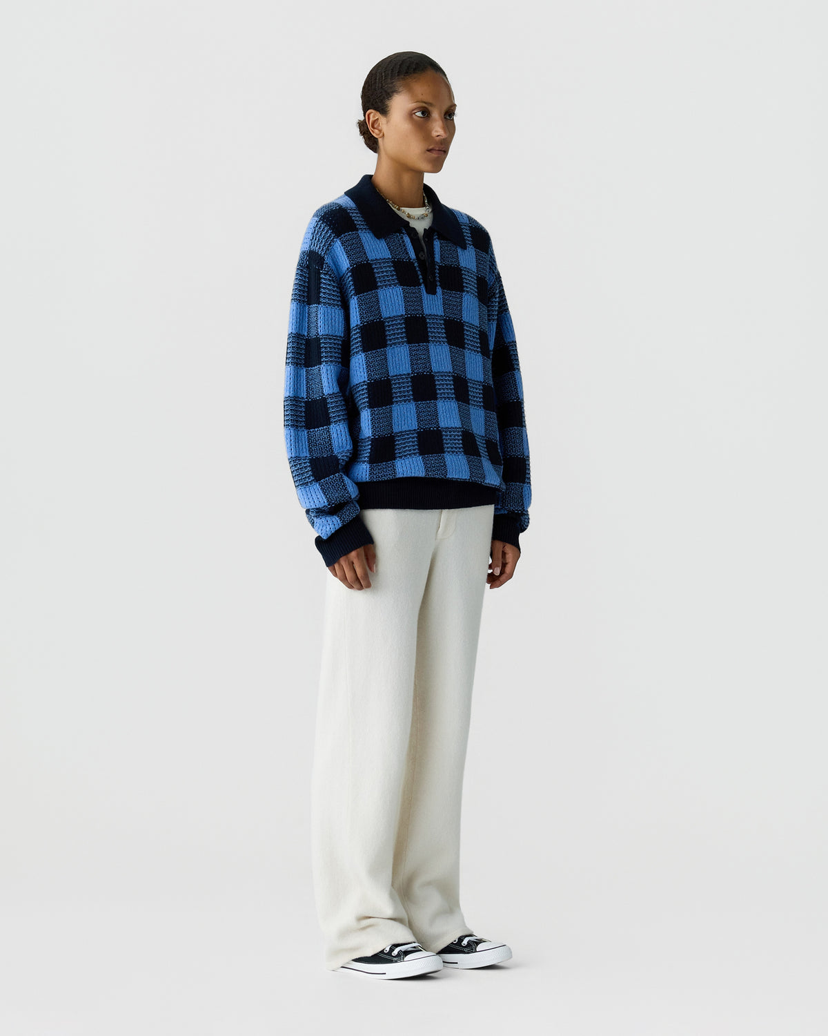 Model wearing a blue and black checkered sweater with white pants and black and white sneakers against a light gray studio background
