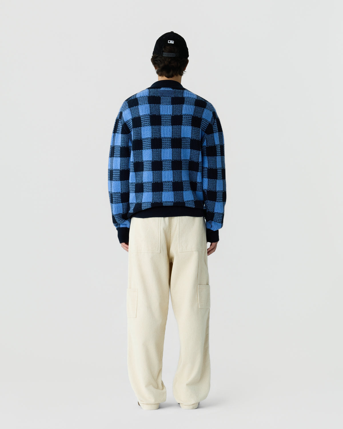 Person wearing a blue and black checkered sweater, cream-colored wide-leg pants, and a black baseball cap, standing facing away against a plain light background.