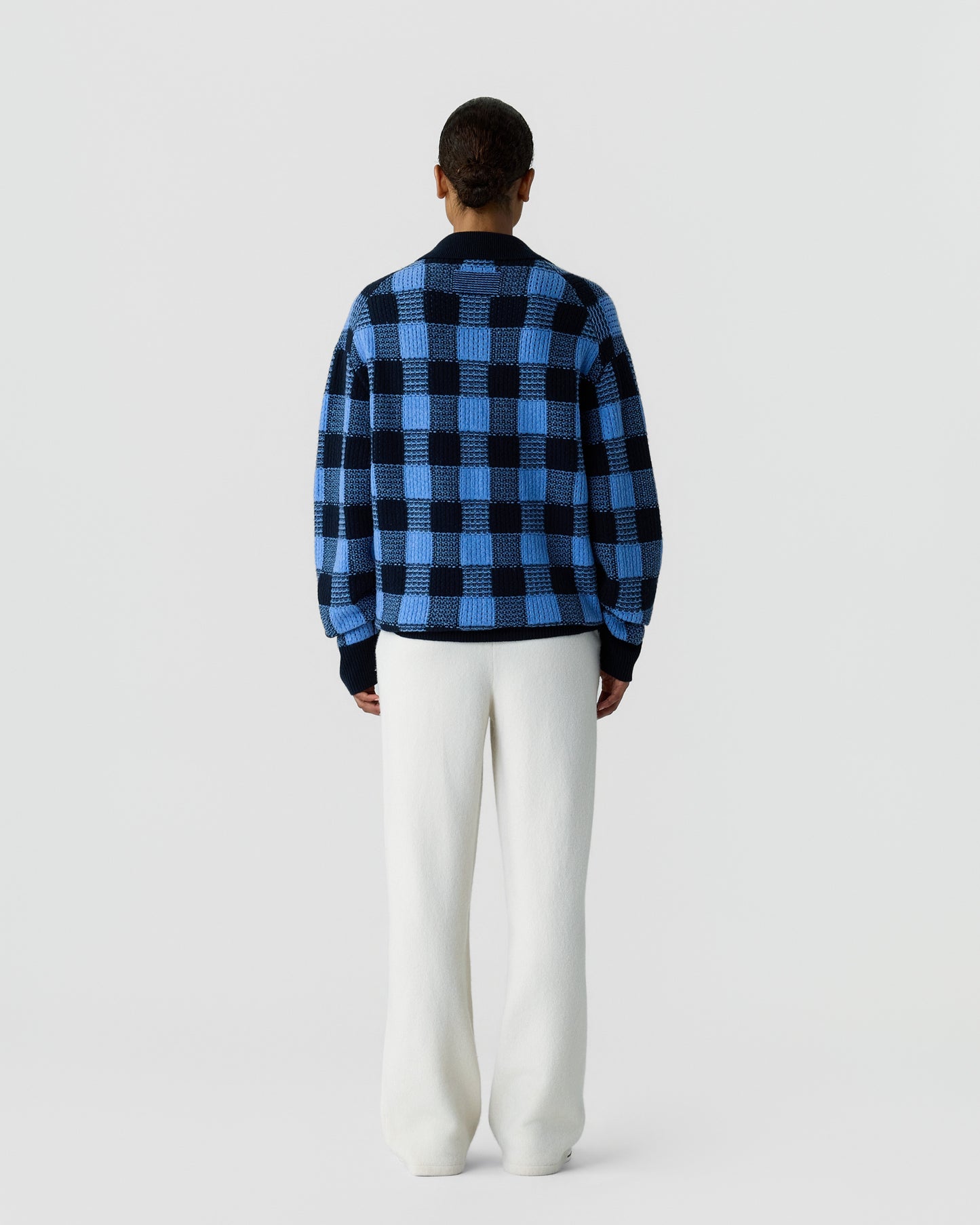 Rear view of a person wearing a blue and black checkered sweater paired with loose-fitting white pants, standing against a plain light gray background.
