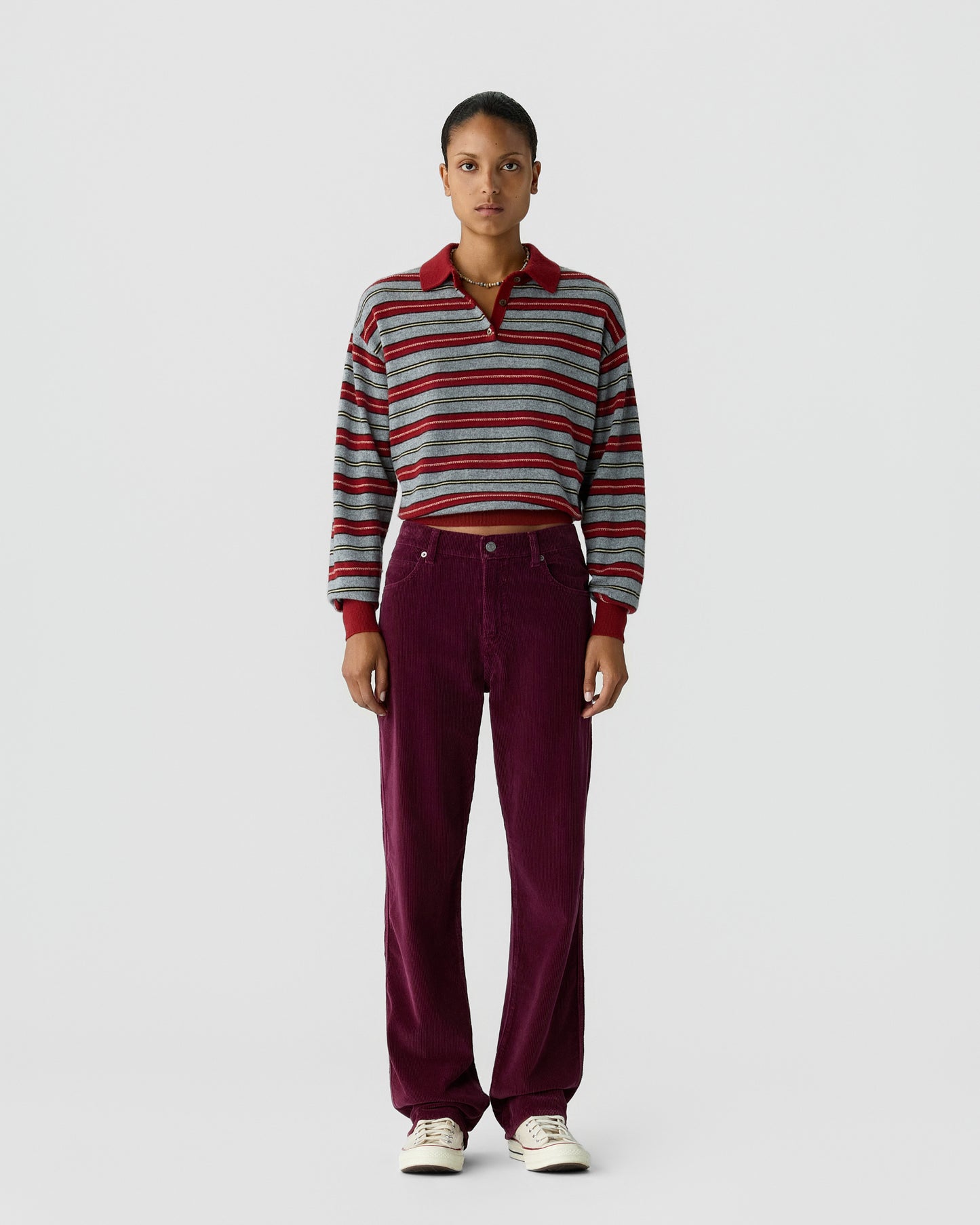 A woman stands against a plain background wearing a striped long-sleeve shirt with a collar and high-waisted, wide-leg purple pants paired with white sneakers.
