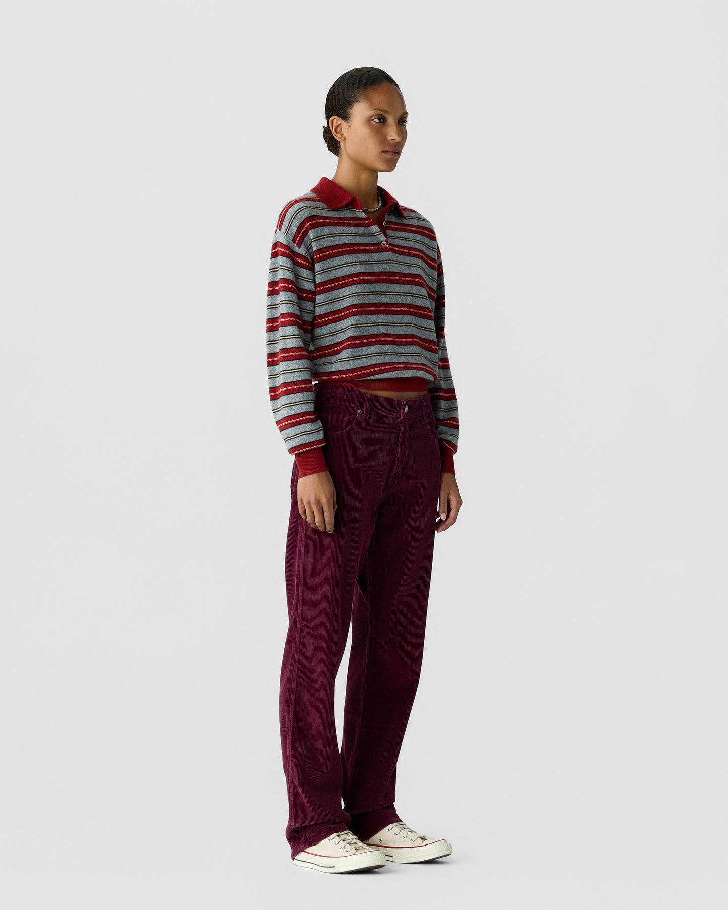 Woman wearing a long-sleeve cropped striped shirt with red collar and cuffs, paired with high-waisted loose-fitting burgundy pants and white sneakers, standing against a plain gray background.