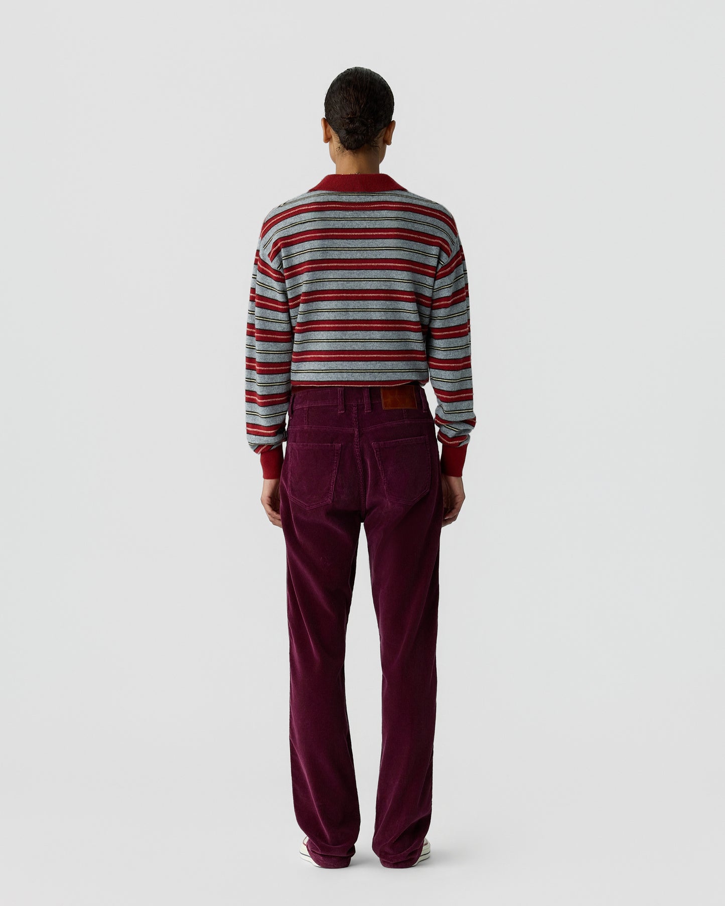 Person standing facing away wearing a cropped, long-sleeve striped sweater and high-waisted, straight-leg burgundy pants against a plain light gray background.