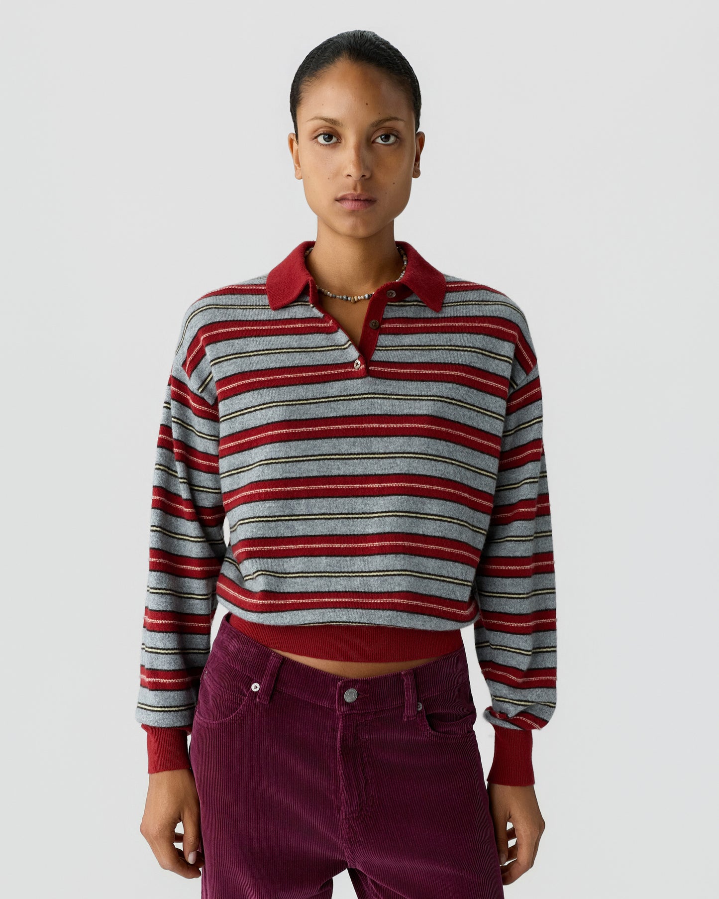 Young woman wearing a long-sleeve striped knit sweater with a red collar and cuffs paired with high-waisted dark red pants against a plain light background.
