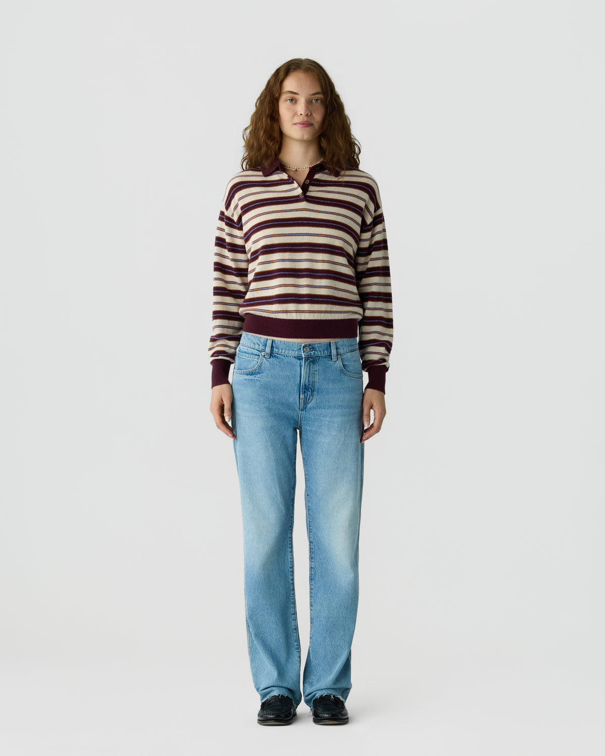 Young woman standing against plain background wearing a striped long-sleeve sweater with maroon and beige tones, light blue jeans, and black shoes