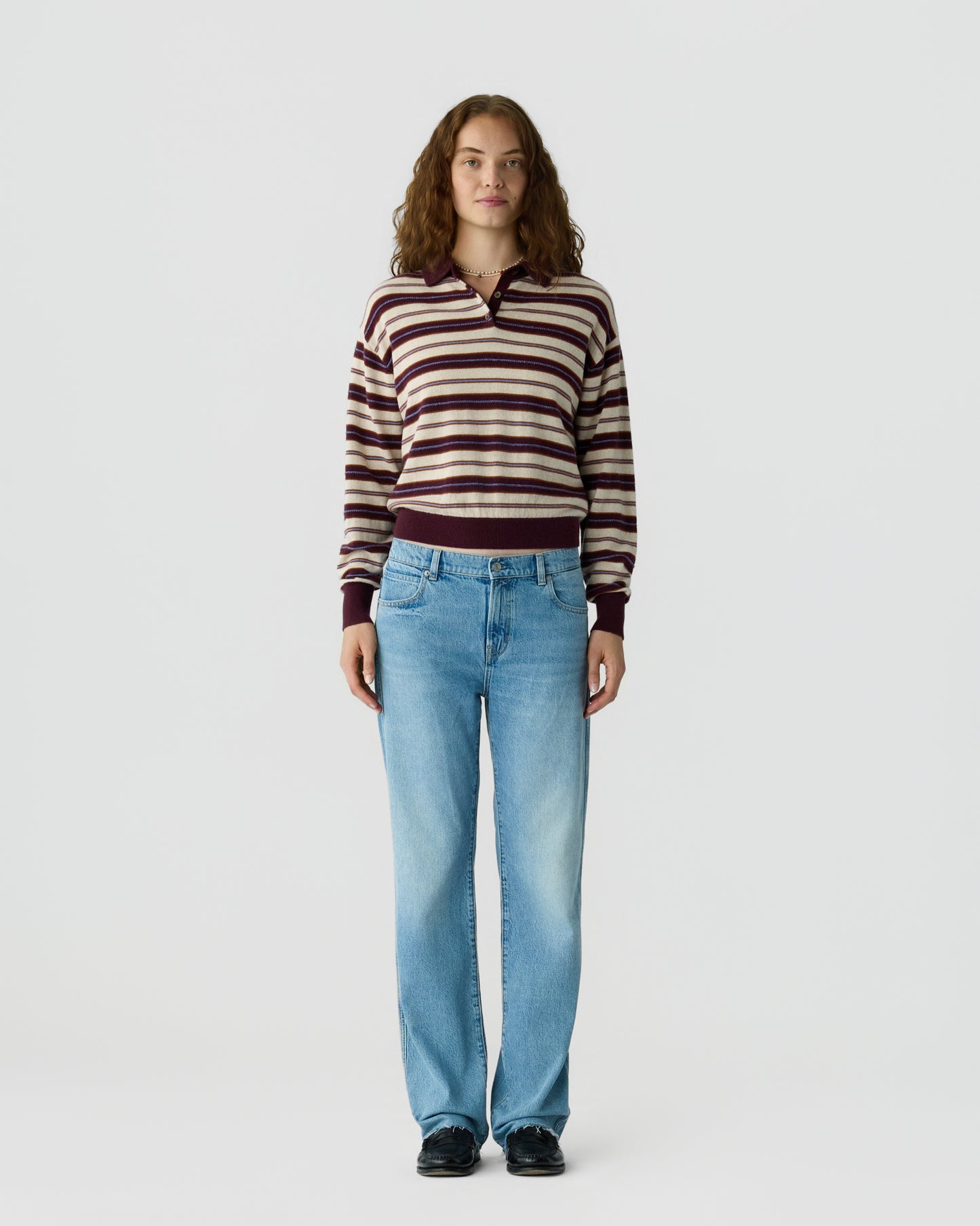 Young woman standing against plain background wearing a striped long-sleeve sweater with maroon and beige tones, light blue jeans, and black shoes