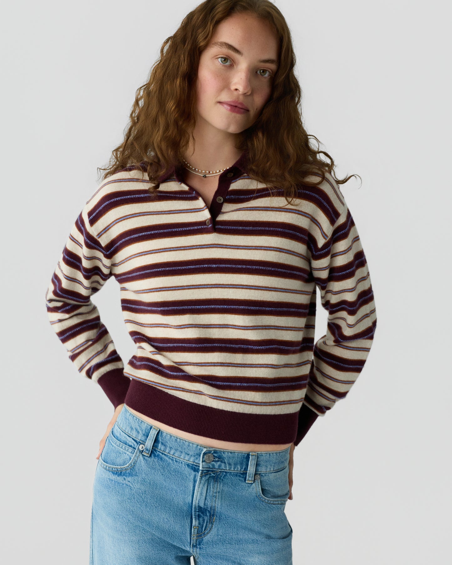 Young woman with curly hair wearing a striped long-sleeve knit sweater and light blue jeans against a plain background